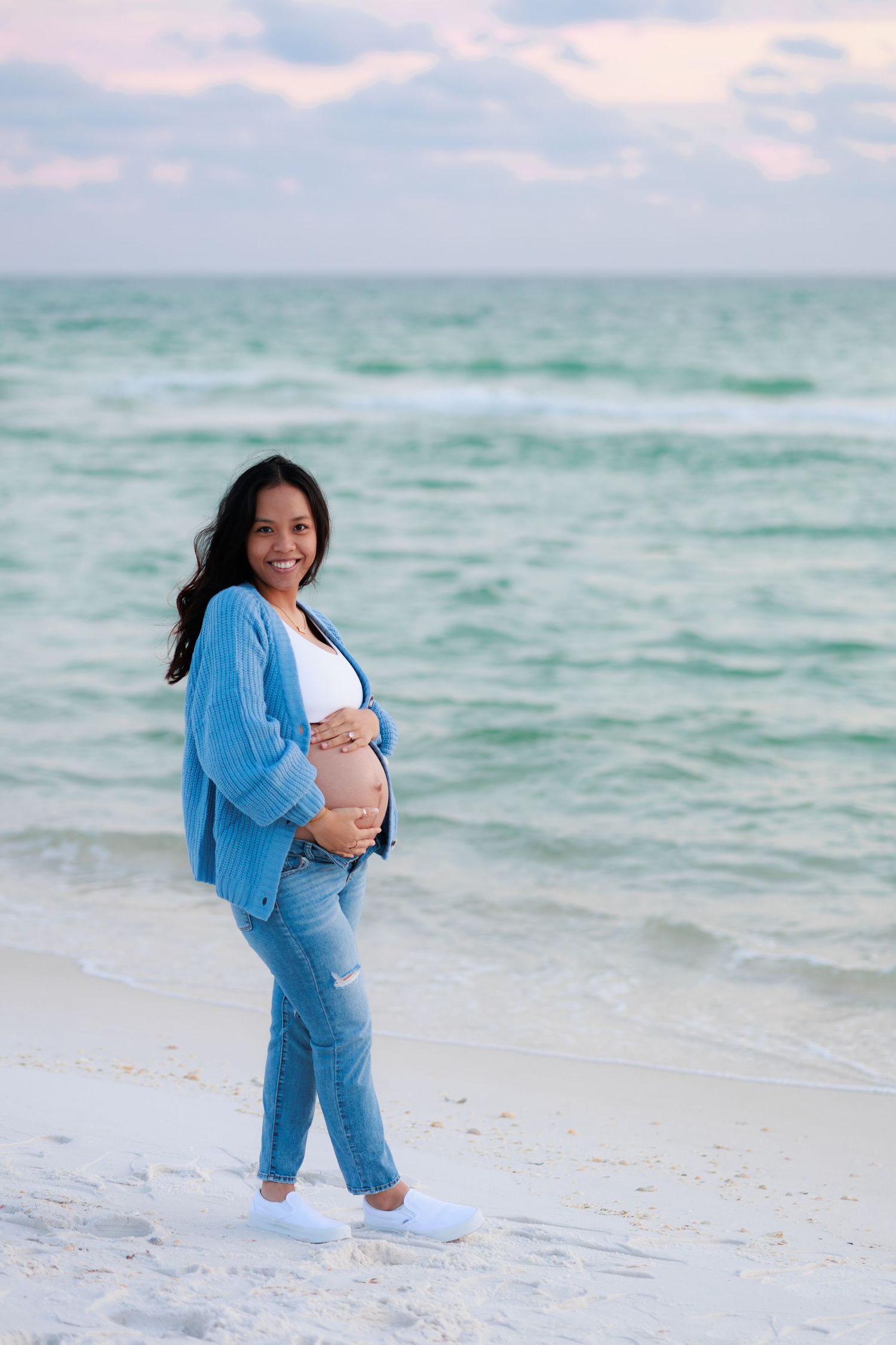 Pregnant woman in blue cardigan poses on beach with ocean waves in background during sunset maternity shoot.