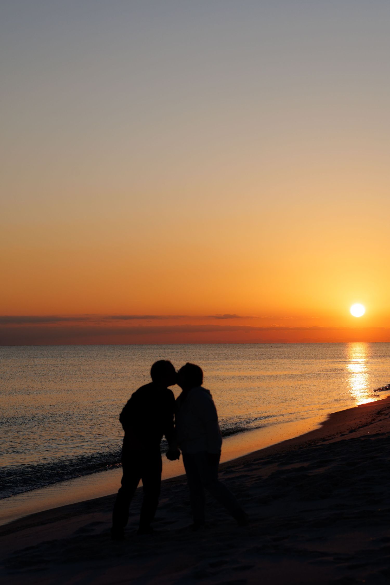 A romantic sunset silhouette sequence shows a couple sitting on rocks by the ocean as the sun descends over the horizon.