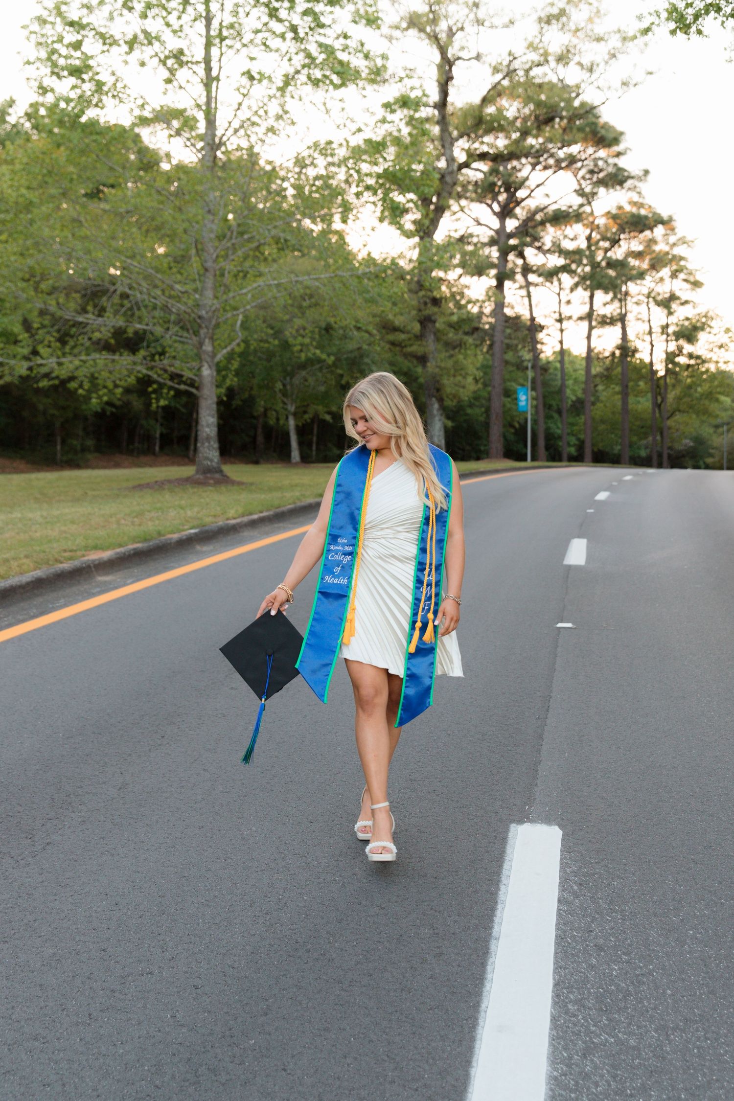 Graduate in blue and white stole tosses graduation cap on empty road surrounded by trees.