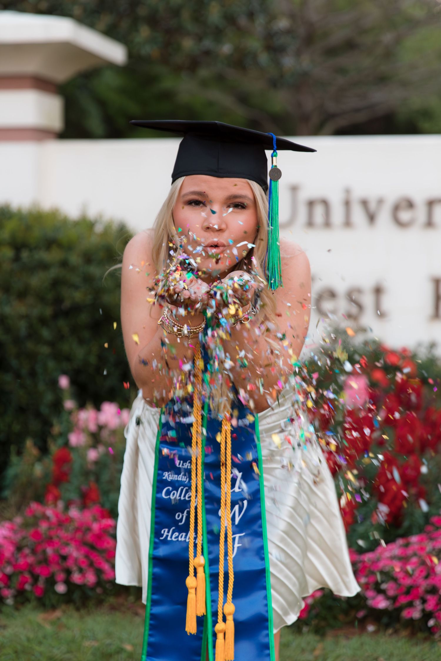 Graduate in cap and gown celebrates by blowing colorful confetti during outdoor graduation ceremony with flowers in background.