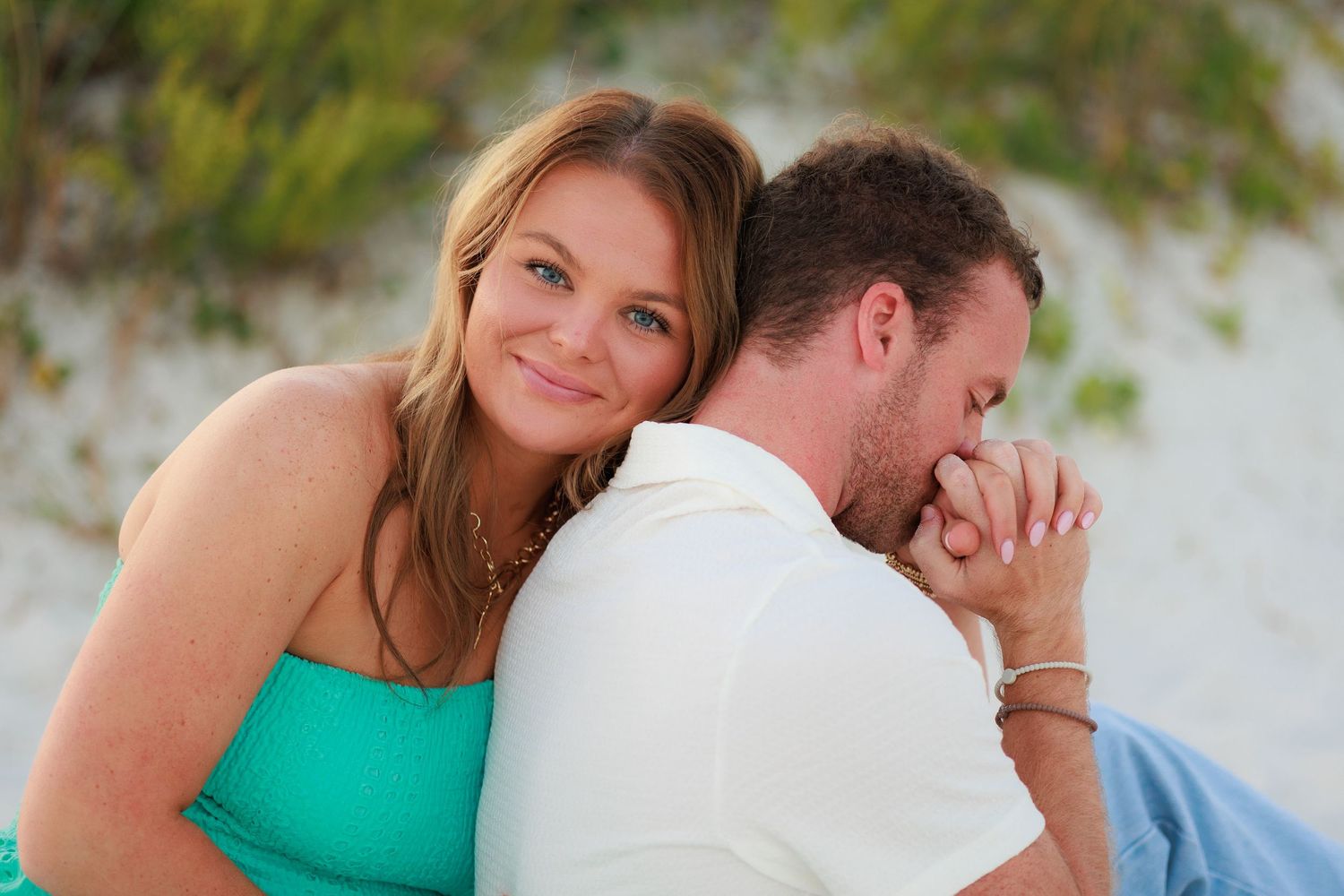 A couple in casual attire shares tender moments together in an outdoor setting with green foliage in the background.
