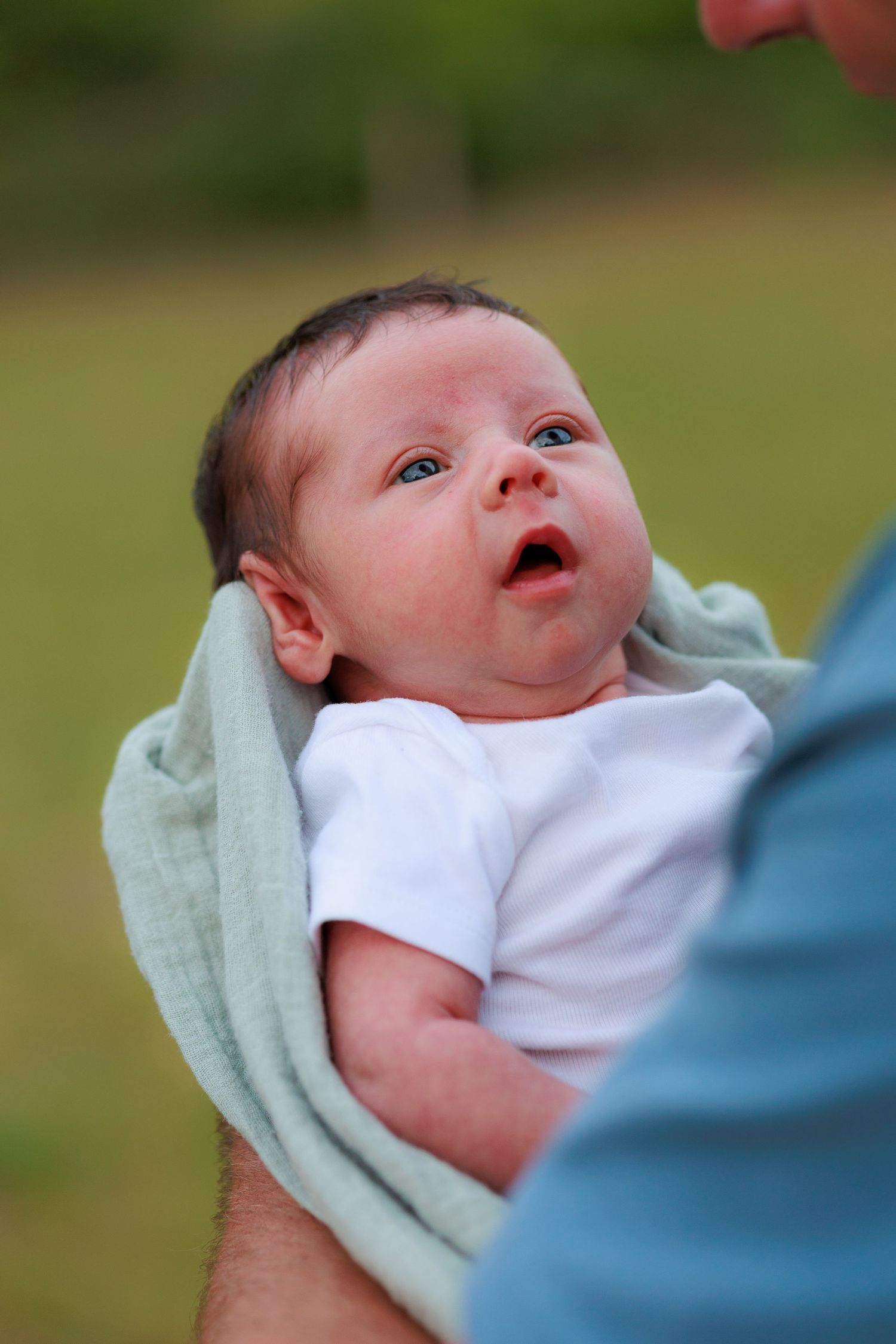 A newborn baby wearing white clothing looks upward while being held in a mint green blanket.