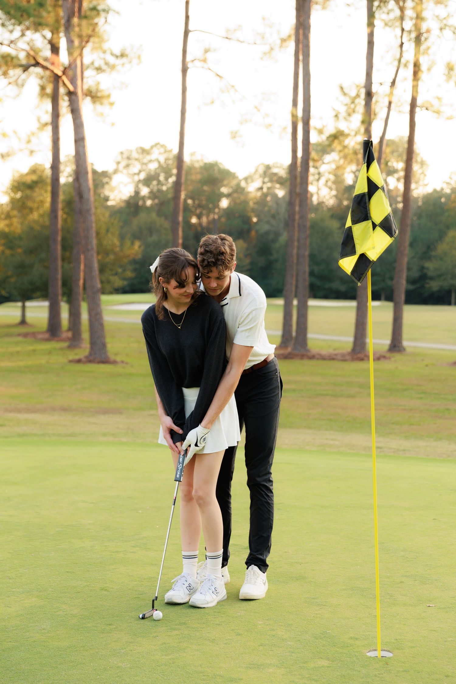 Two golfers practice putting techniques together on a sunny golf course green with tall pine trees in the background.