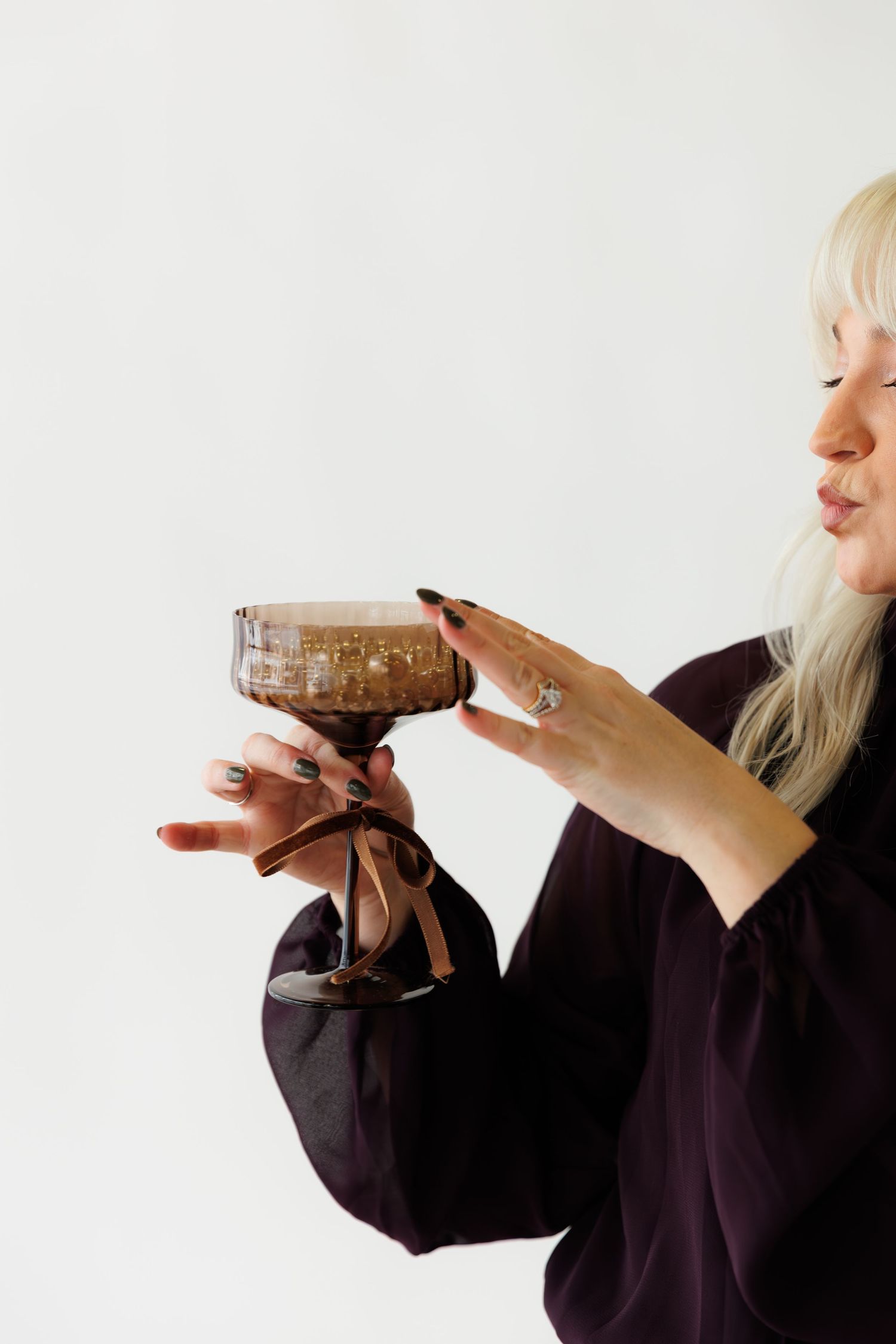 Person in black clothing holds and sips from a fancy cocktail glass against white background.