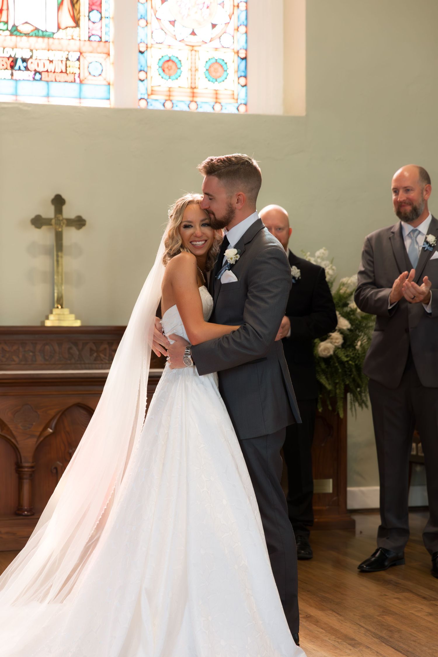 Newlyweds embrace during their ceremony in a sunlit church with stained glass windows.