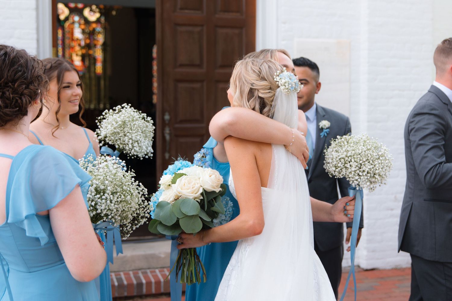 Wedding party gathered outside a church entrance with bridesmaids in light blue dresses holding bouquets.