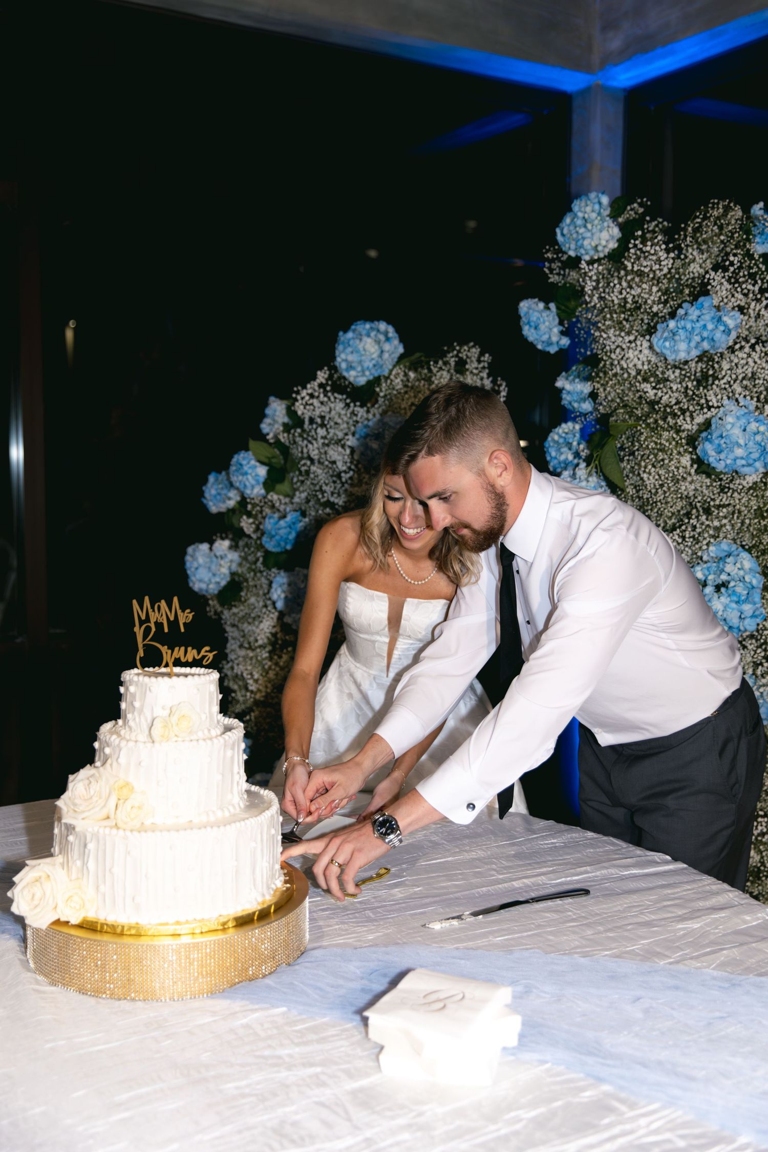 Newlyweds cutting their elegant white wedding cake together in front of blue floral decor.