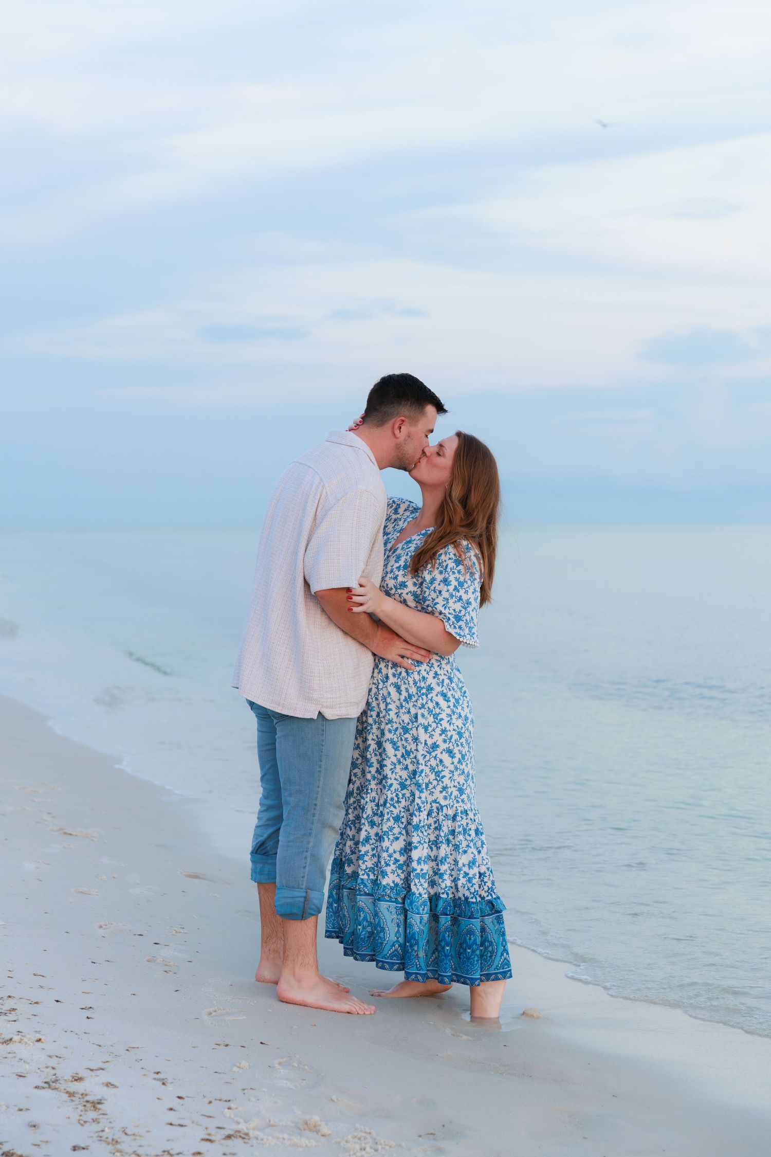 A couple shares a romantic kiss while embracing barefoot at the water's edge on a white sand beach at dusk.