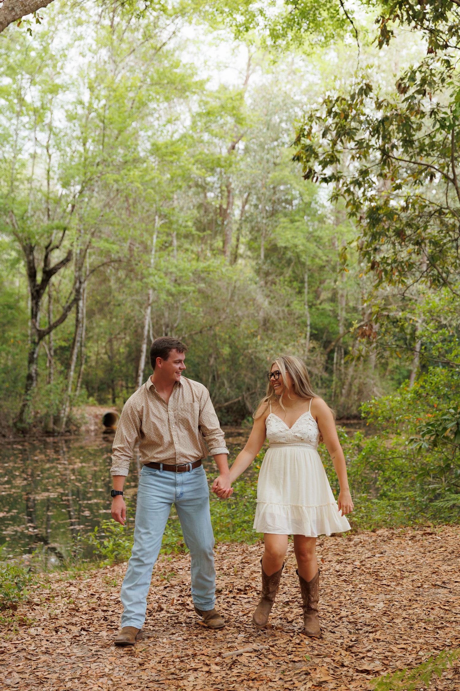 Couple holding hands walking through a lush forest path, man in jeans and woman in white dress with cowboy boots.