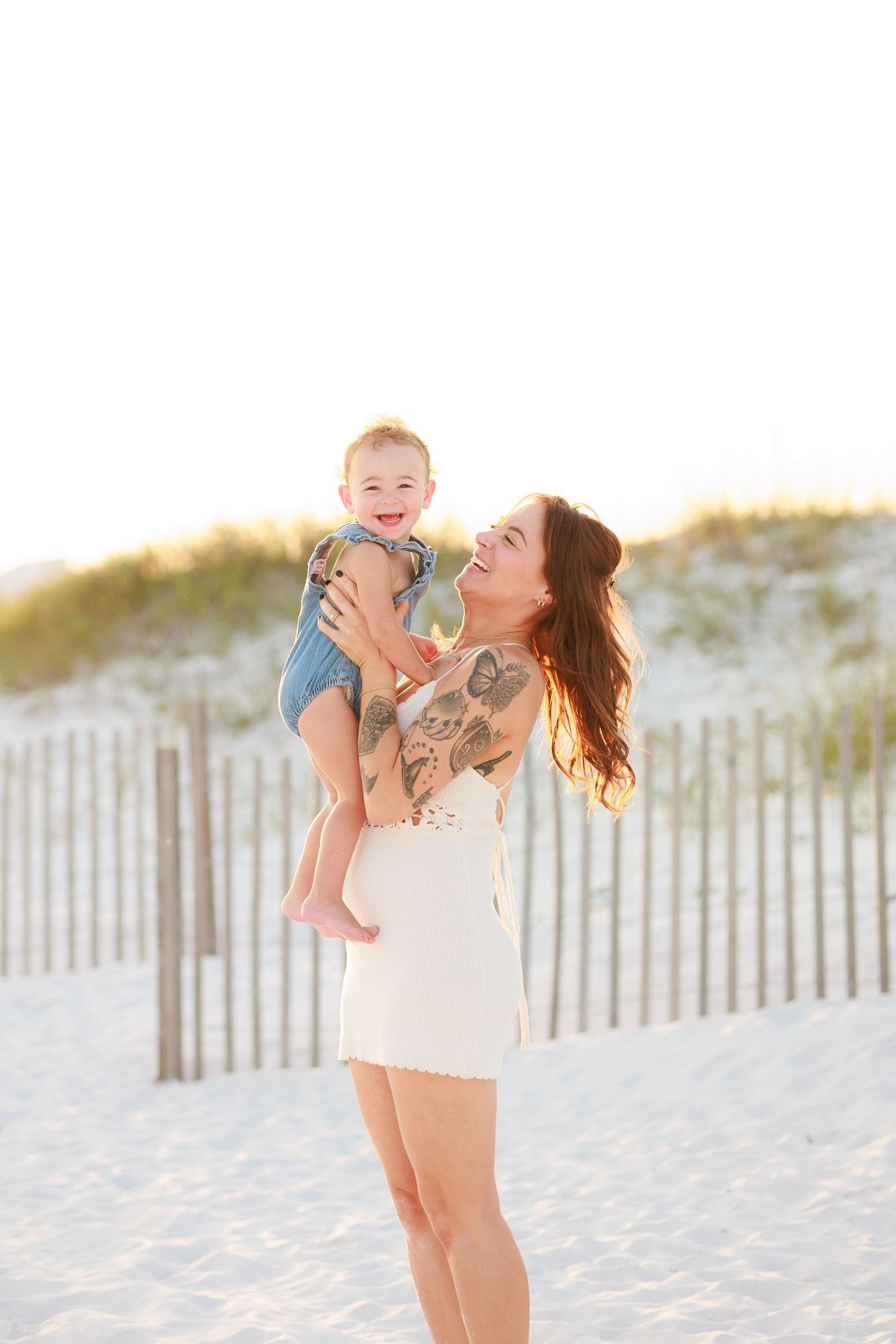 Tattooed mother in white dress lifting laughing baby on sandy beach with dunes.