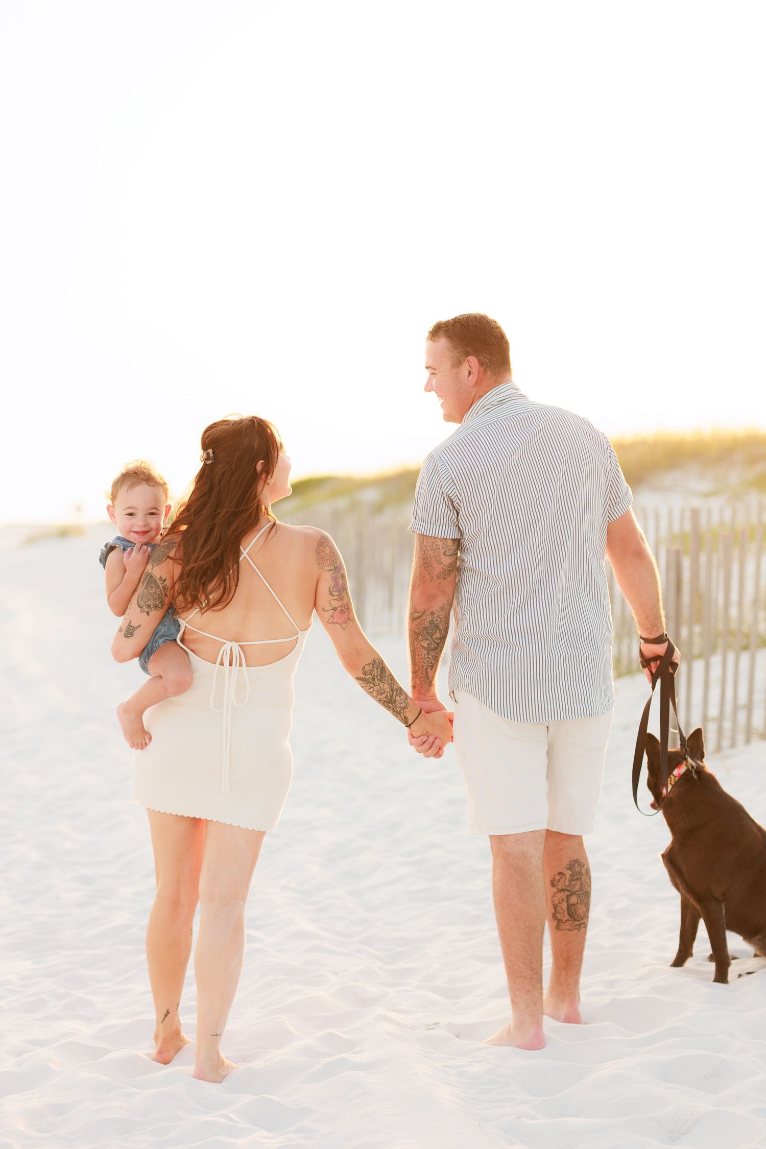 Young family holding hands walking on white sand beach, woman carrying baby, man leading dark dog at sunset.