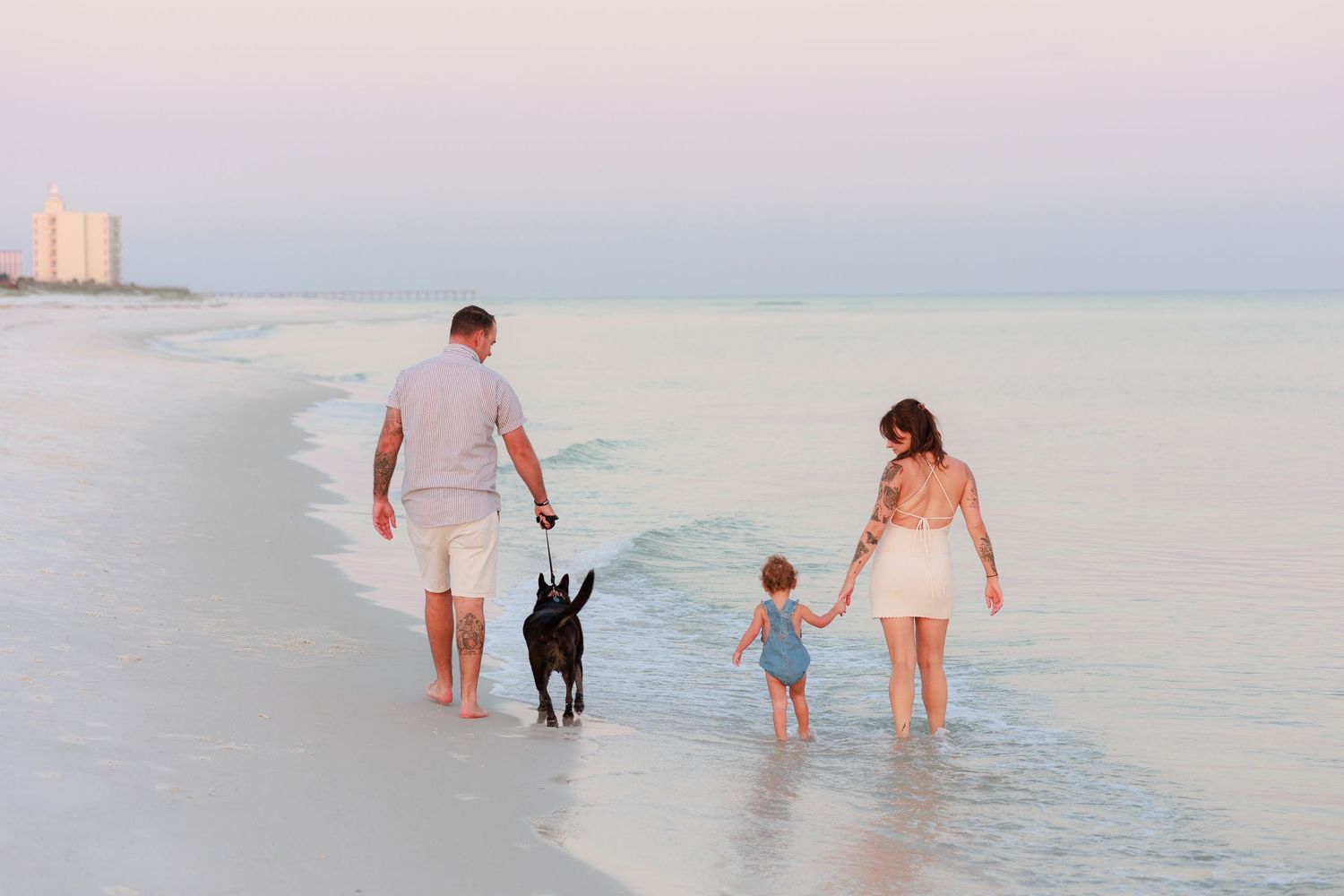 Family with toddler and black dog walking along a serene beach at sunset, waves gently lapping the shore.
