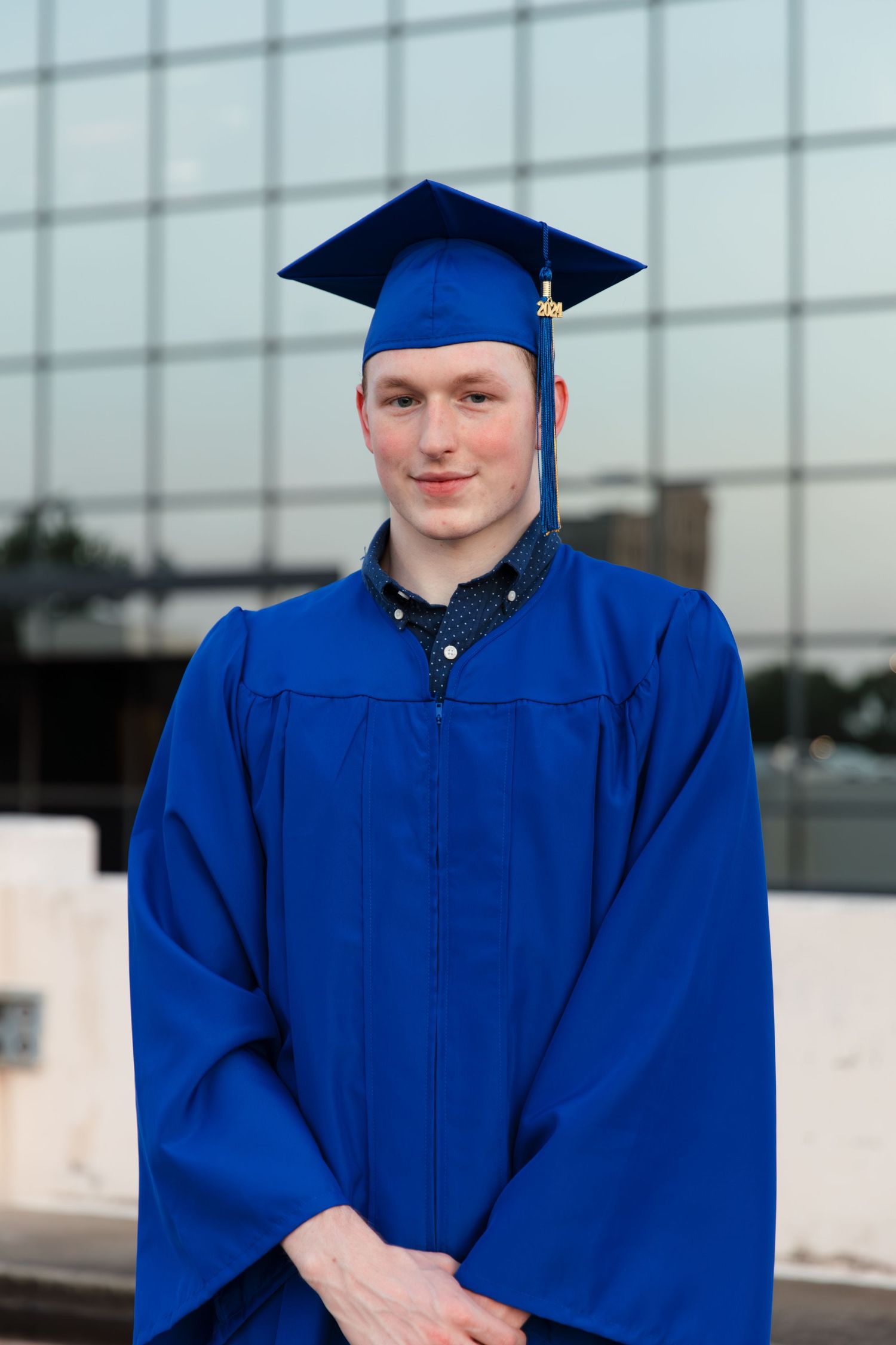 Young man in blue graduation cap and gown smiling outdoors in front of a modern glass building.