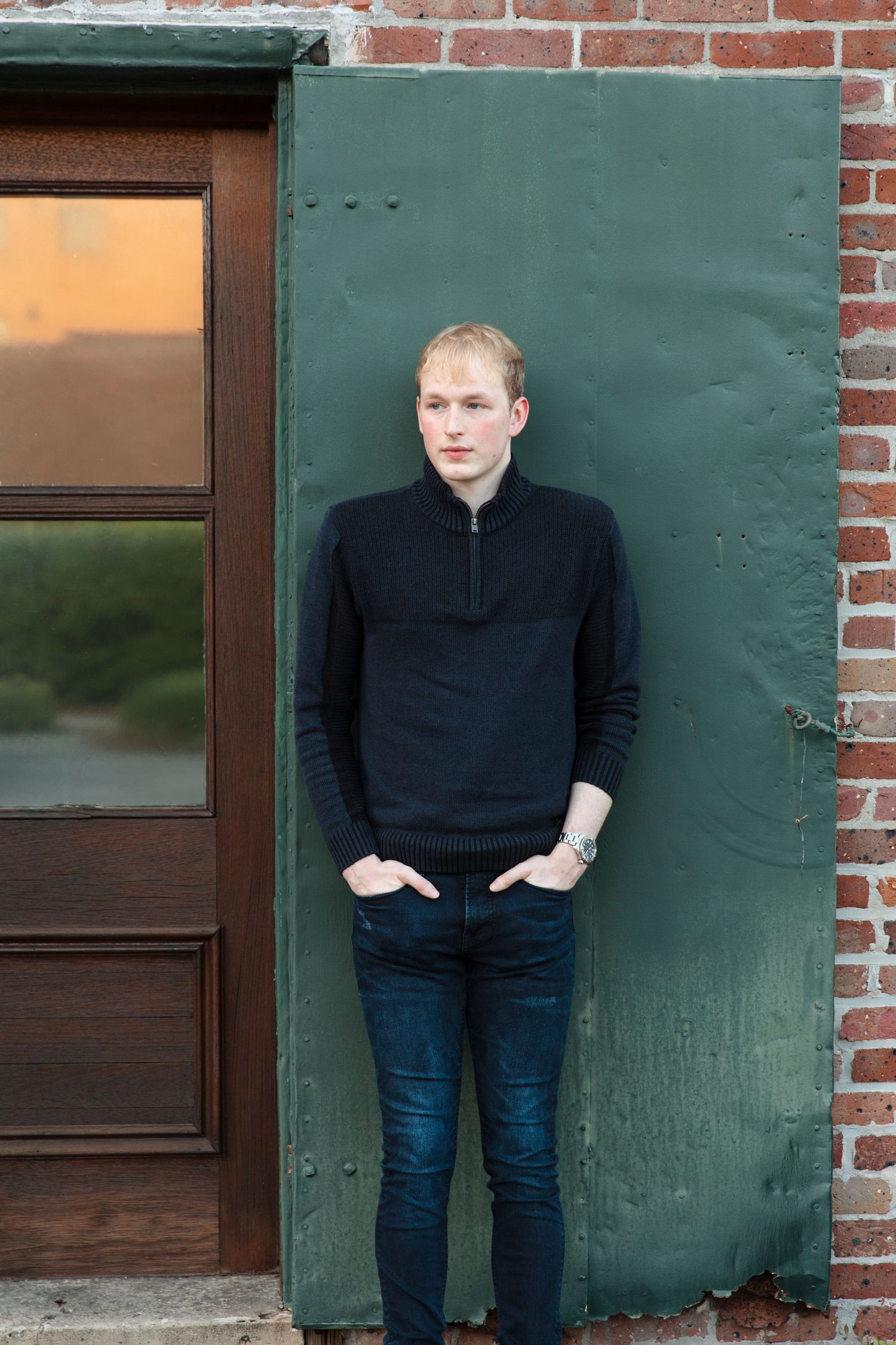 Young man in black sweater and jeans leaning against a green metal wall beside a wooden door.