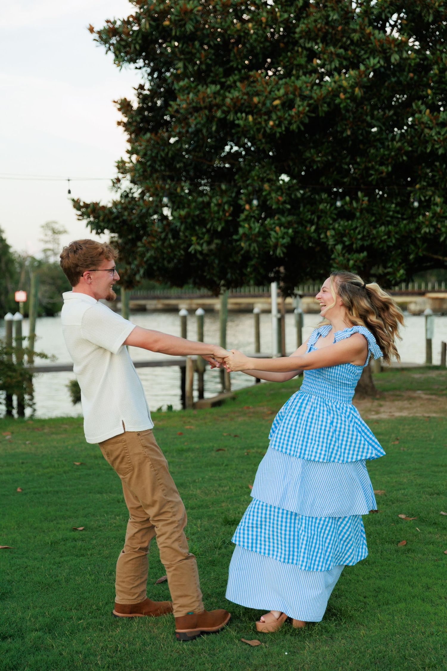 A happy couple dances and spins together on a green lawn near a waterfront dock surrounded by lush trees.