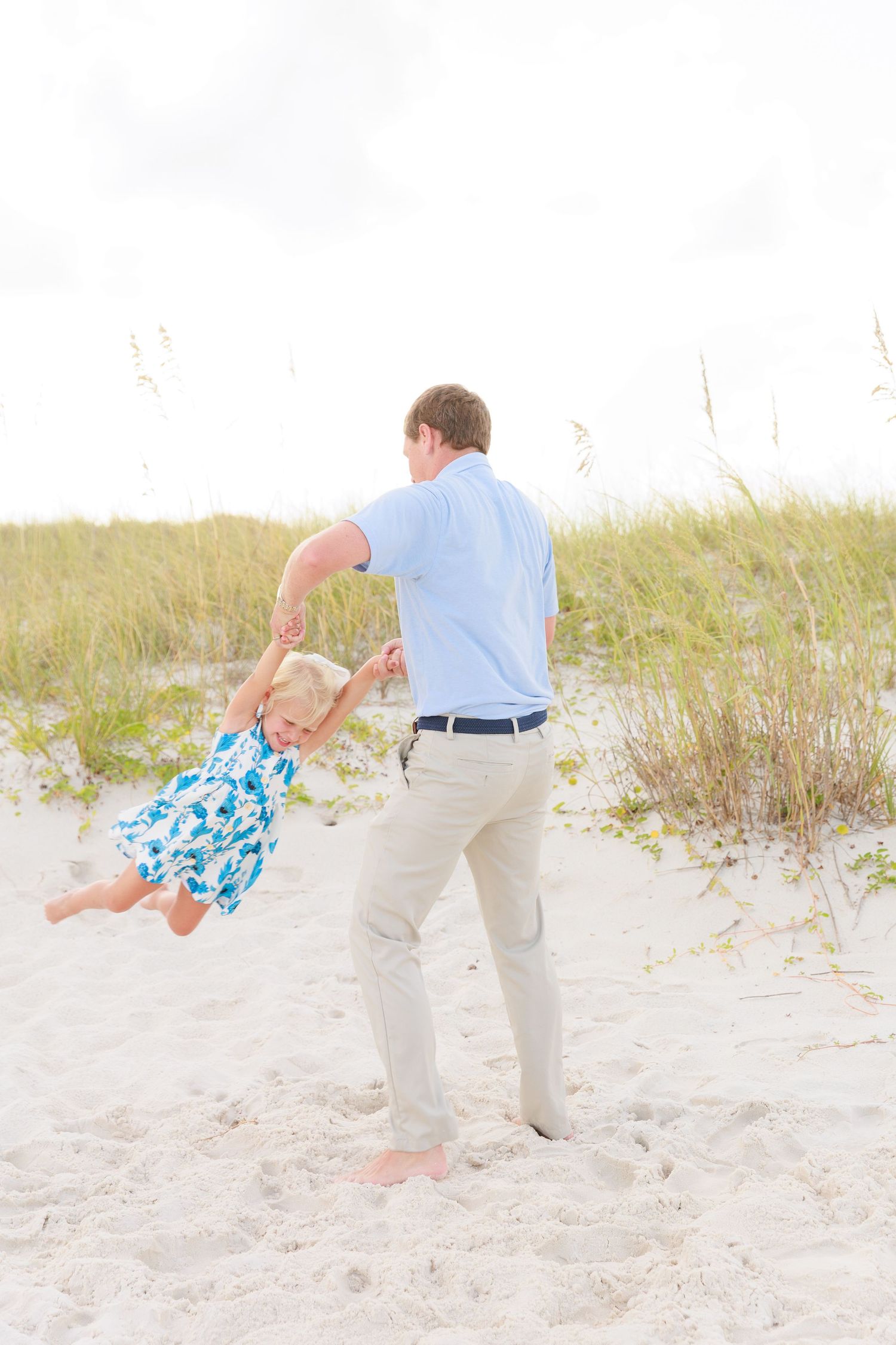 A father swings his young blonde daughter in a blue floral dress on a white sand beach near sea oats.