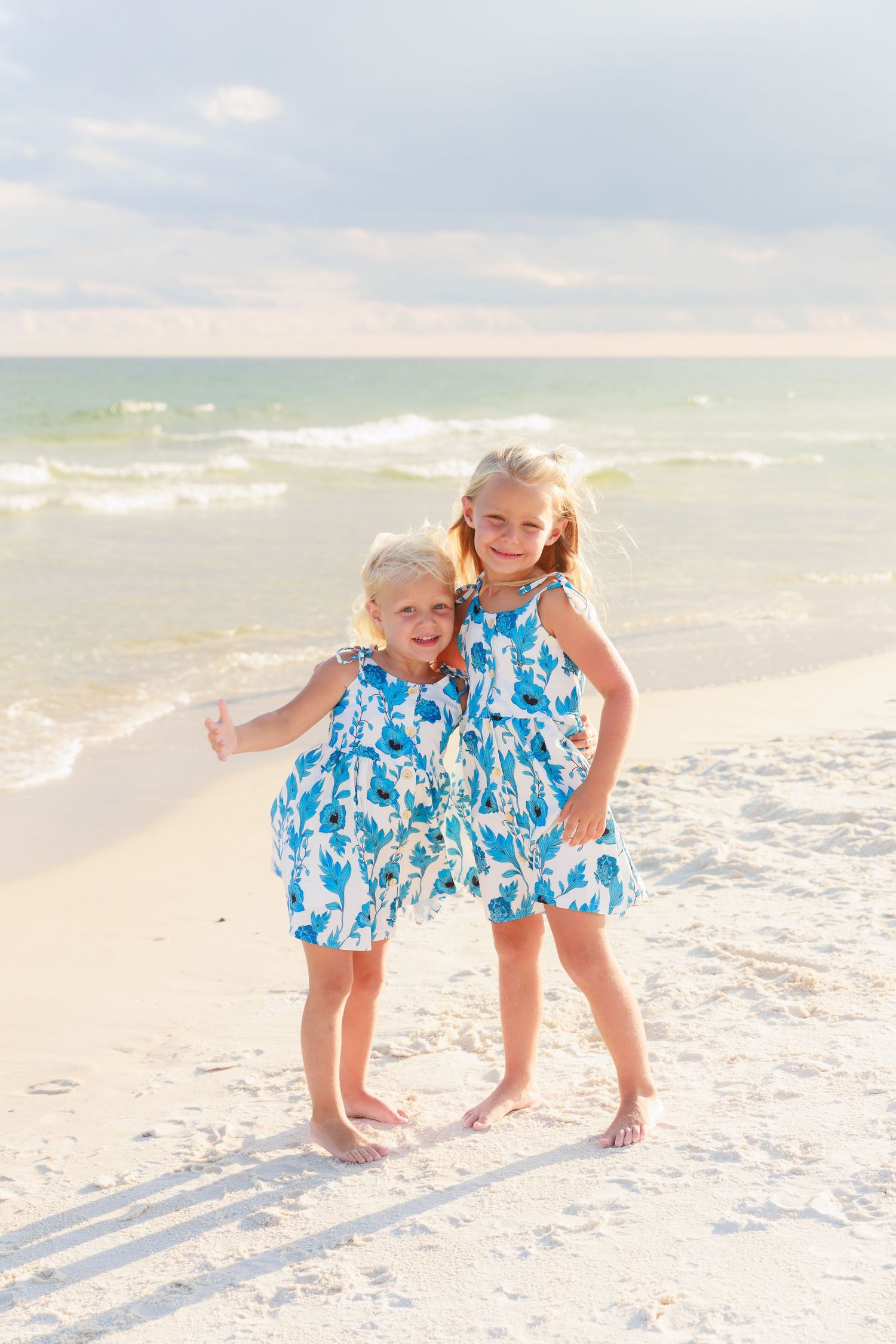 Two young sisters in matching blue floral dresses hugging and smiling on a sunny white sand beach.