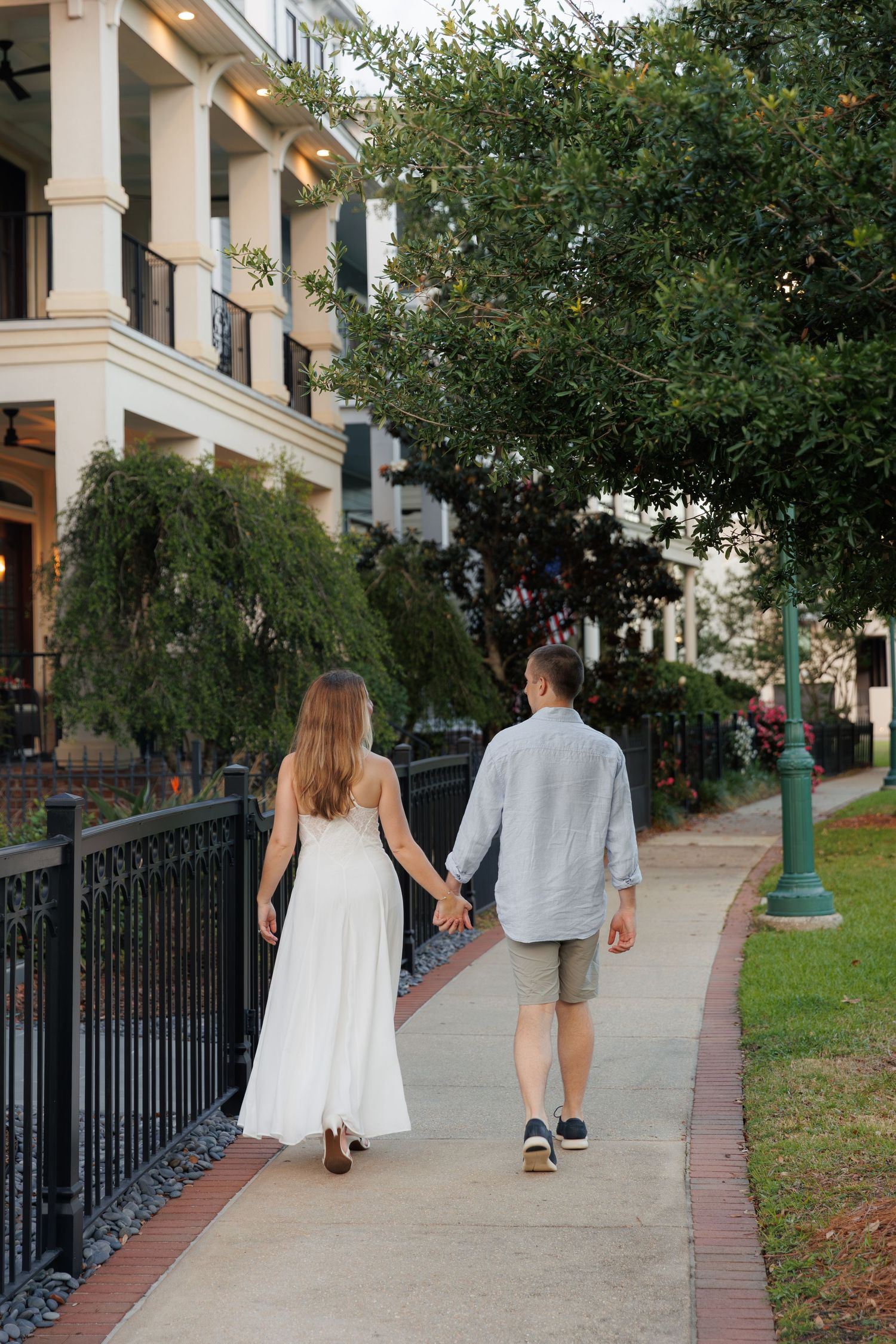 Couple holding hands walking down a tree-lined sidewalk, woman in white dress and man in gray shirt and shorts.