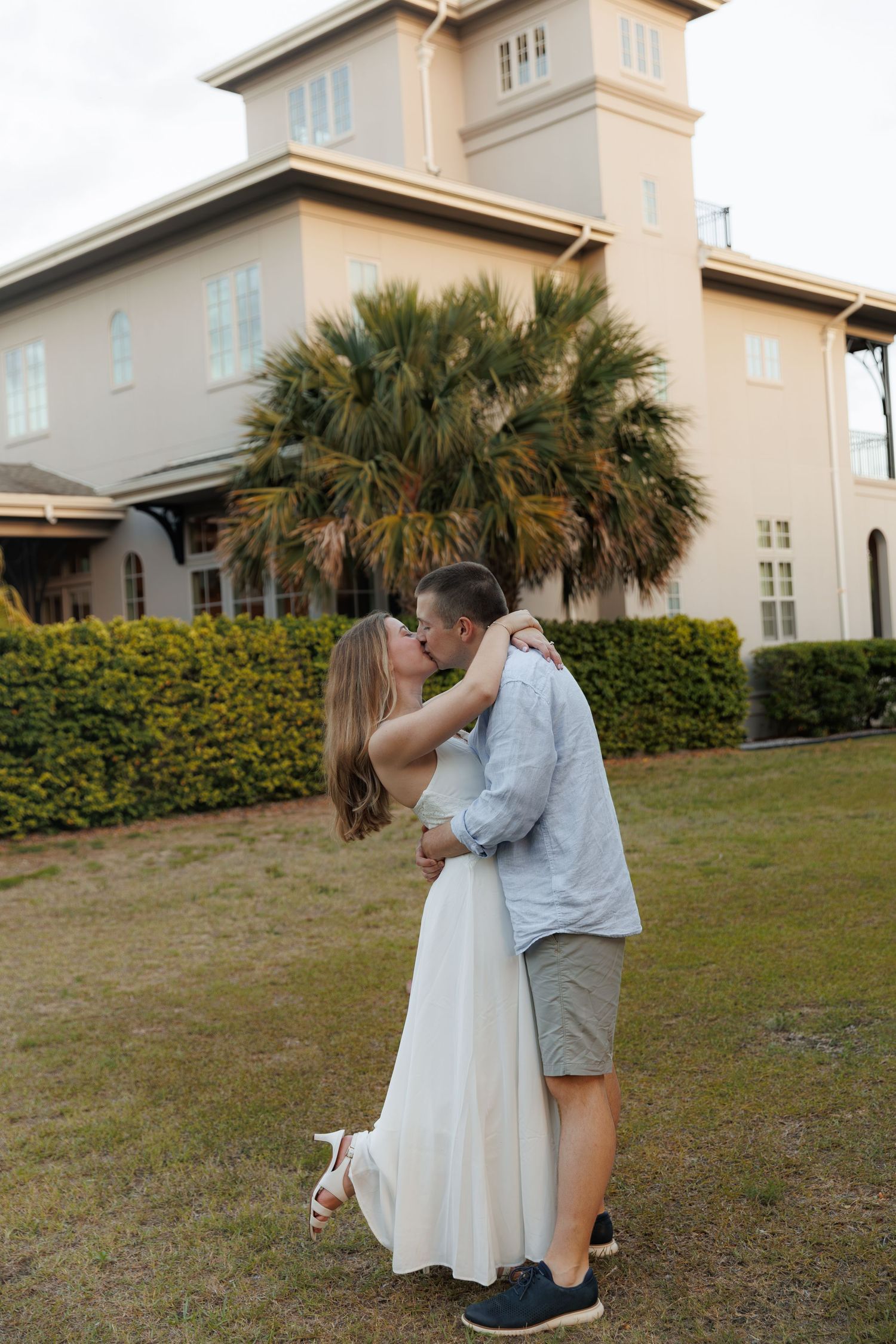 Couple sharing a romantic kiss on green lawn with large palm tree and beige building in background.