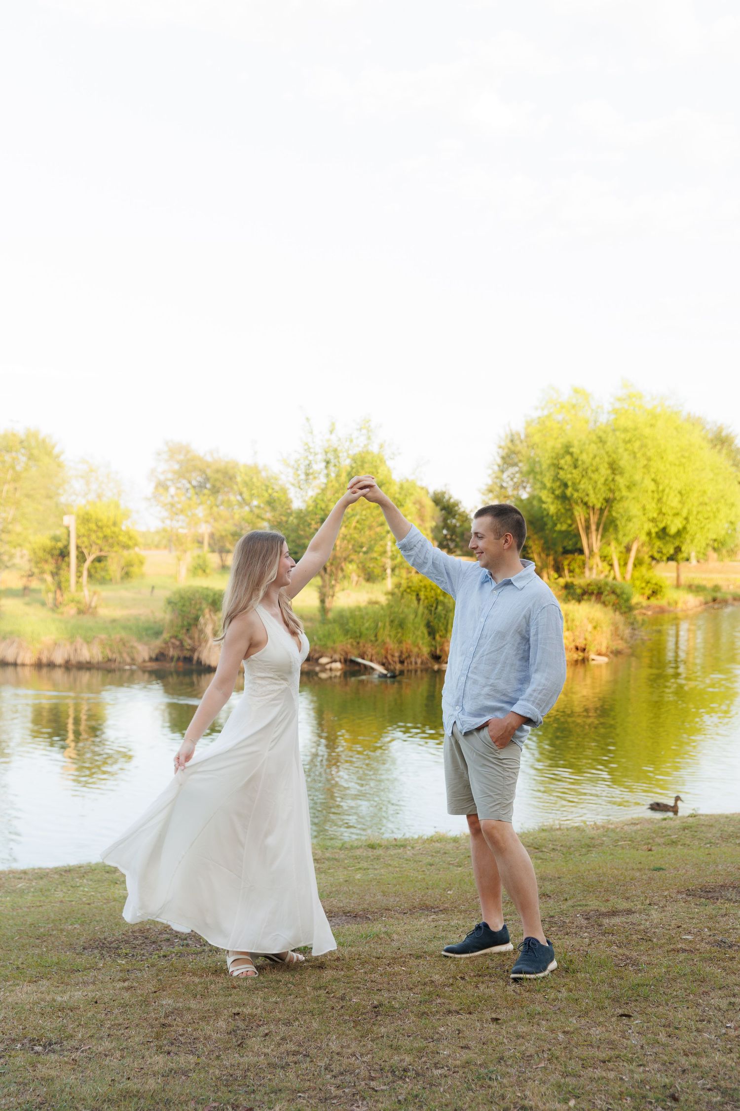 Couple dancing by a sunlit pond, woman in white dress twirling, man in blue shirt and shorts, lush green trees background.
