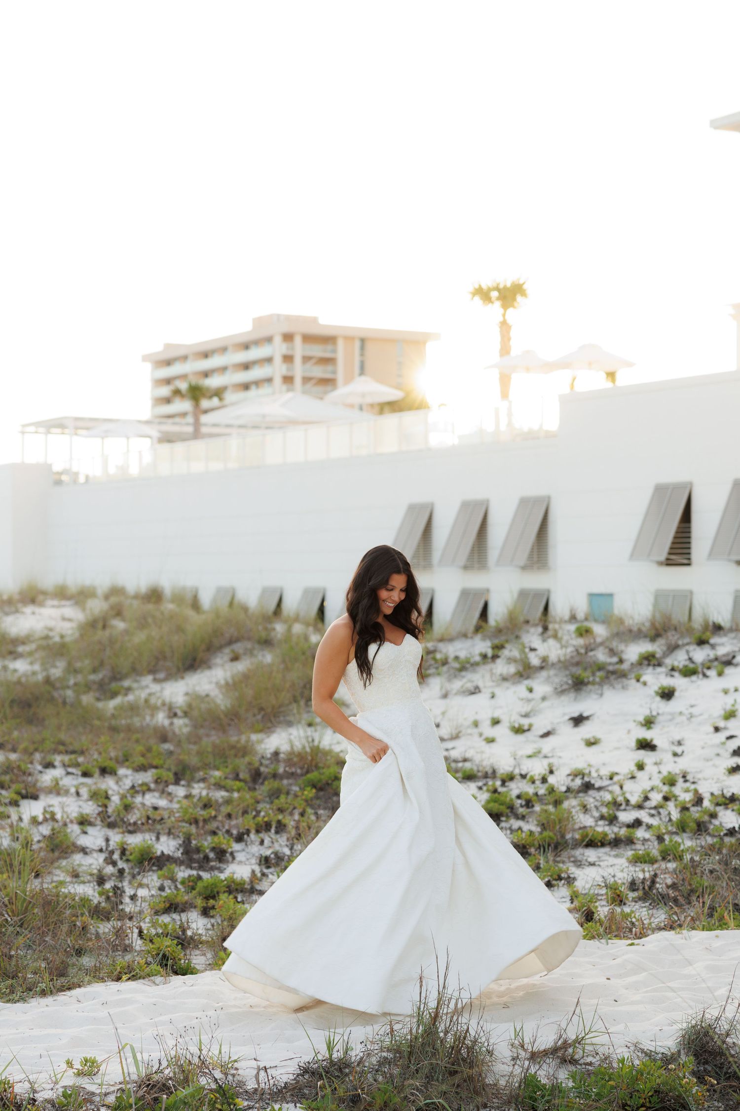 Bride in white ball gown twirling on sandy beach with coastal building backdrop in soft golden light.