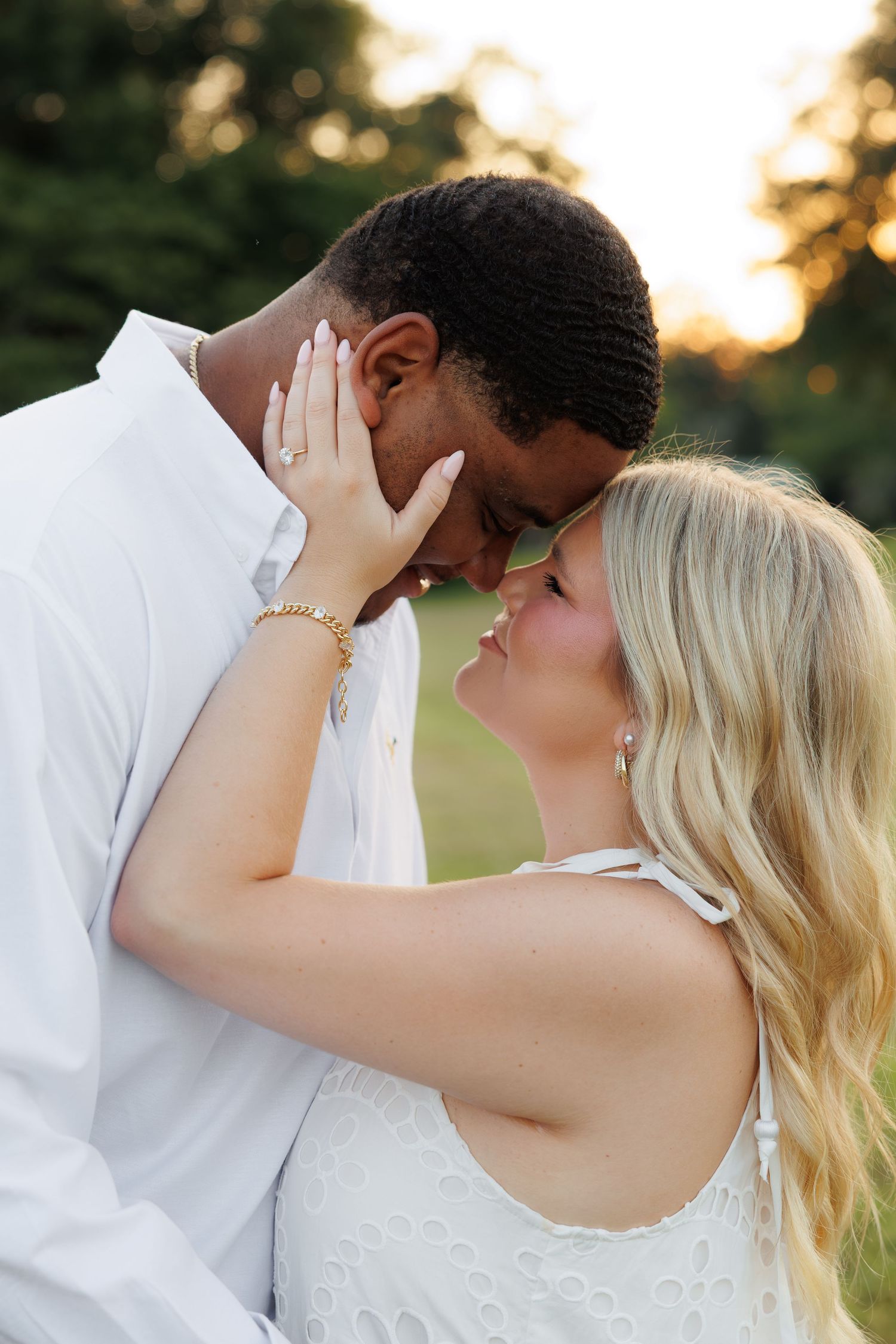 Couple in white outfits sharing a romantic moment outdoors at sunset, woman showing engagement ring.