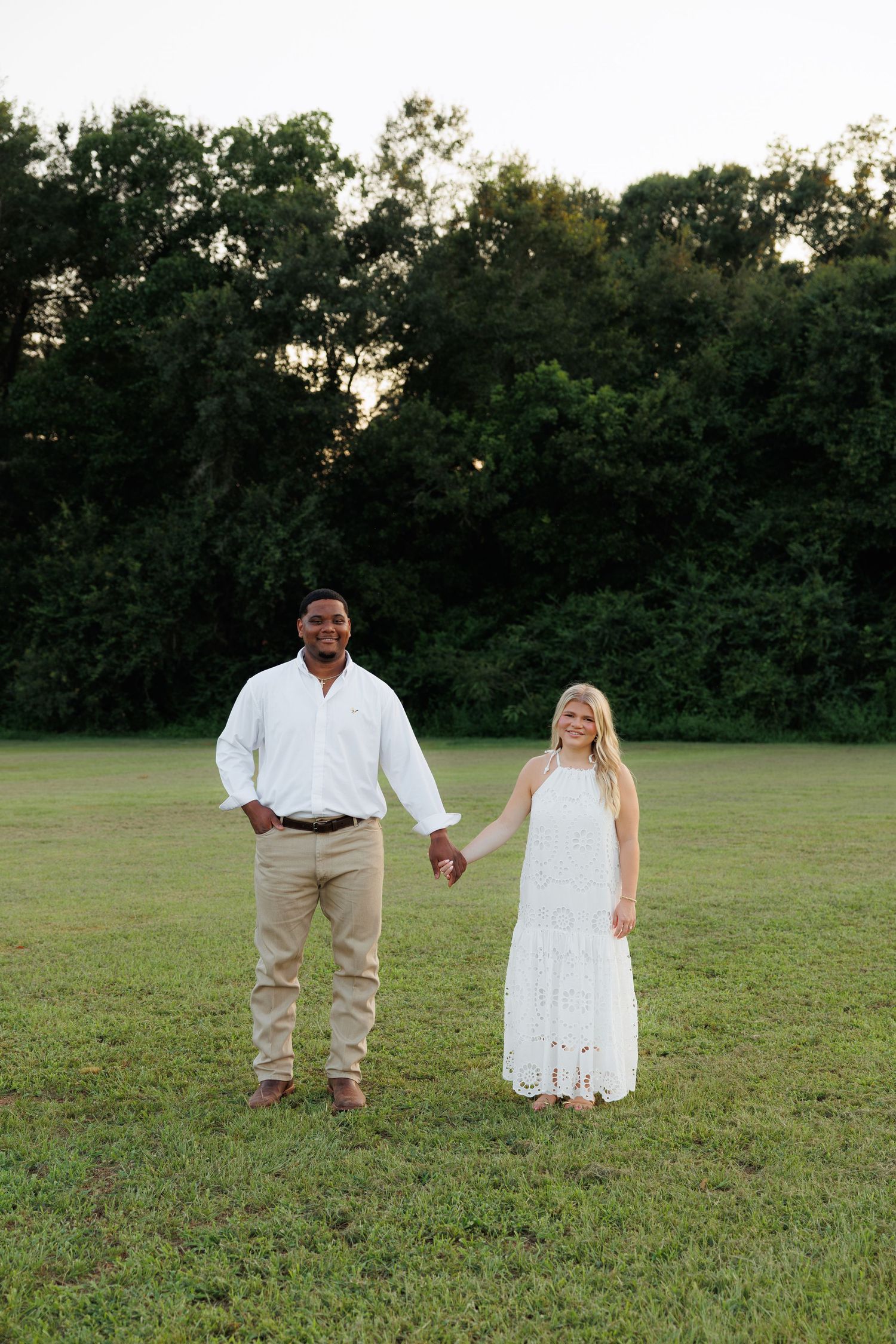 A couple holds hands in a green field at sunset, man in white shirt and khakis, woman in white lace maxi dress.