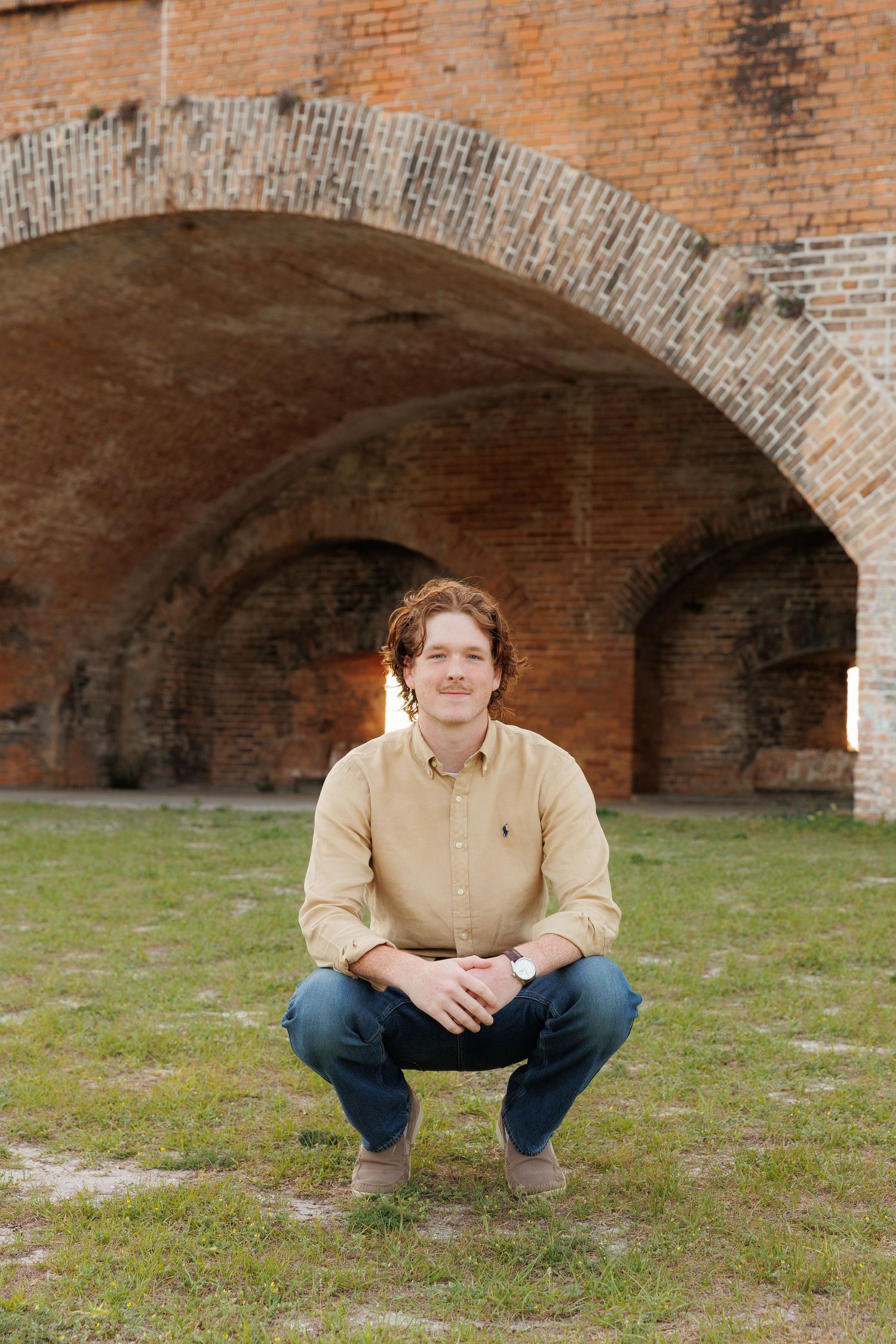 Man in beige shirt crouching on grass before a rustic brick archway, smiling confidently outdoors.