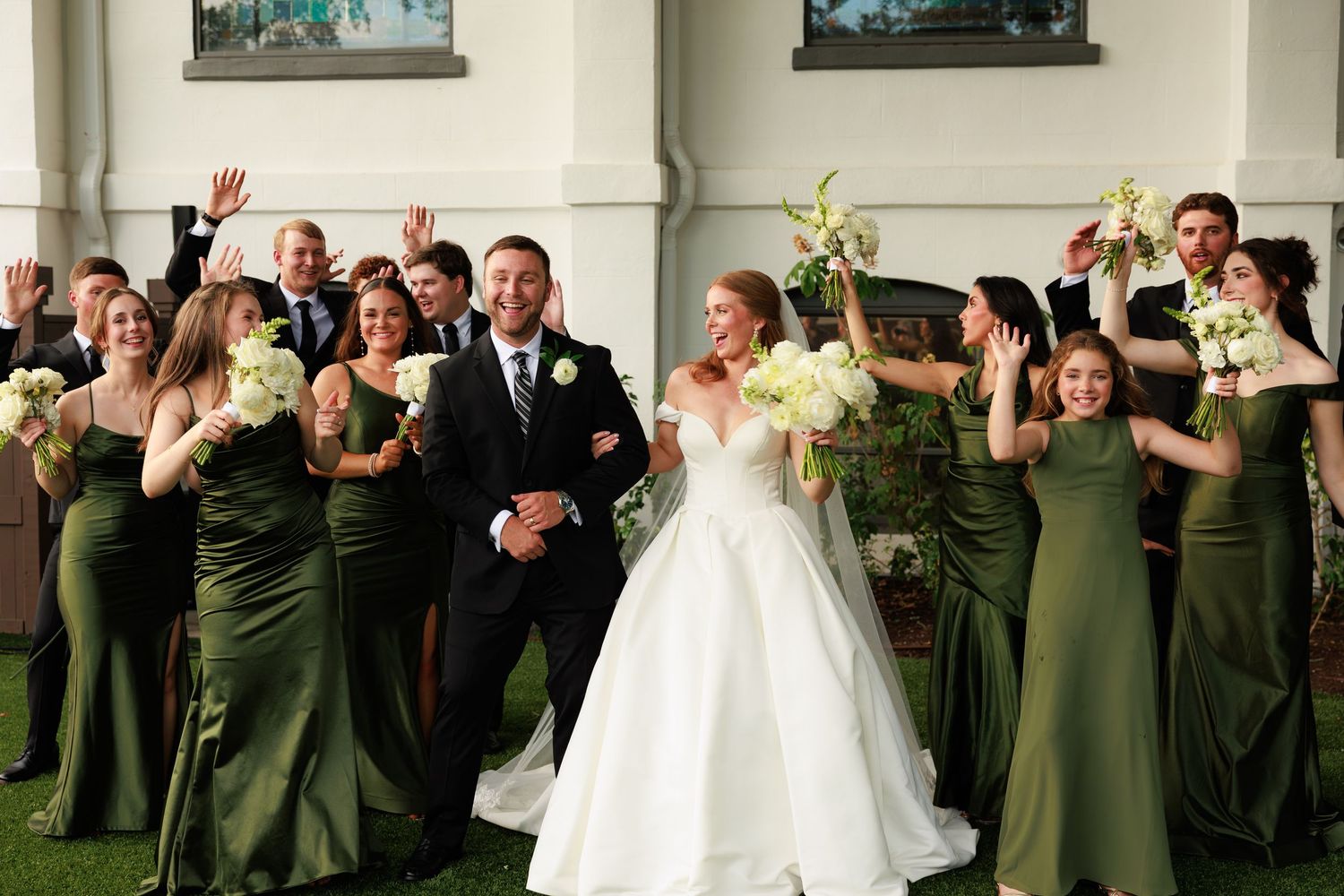 Bride and groom celebrate with wedding party in olive green bridesmaid dresses, raising arms joyfully outdoors.