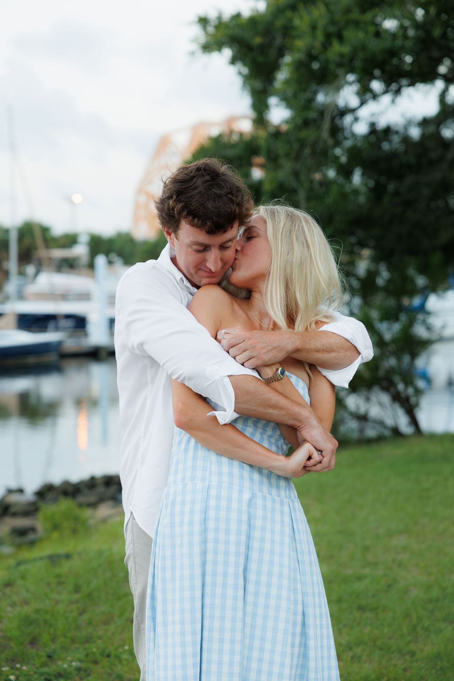 Loving couple shares a tender embrace and kiss on the cheek by a marina.