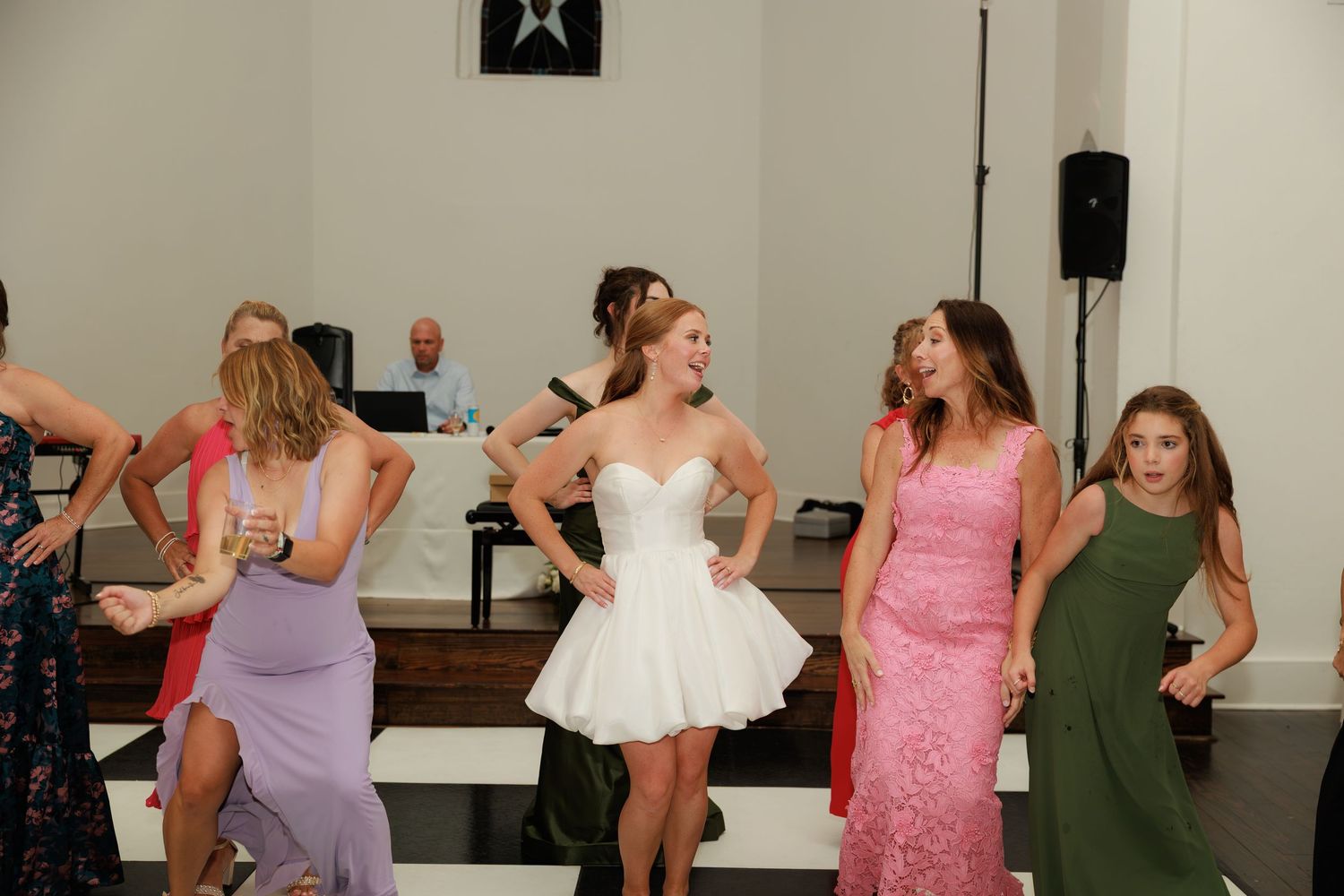 Bridesmaids in colorful formal gowns dancing together at a wedding reception on a white dance floor.
