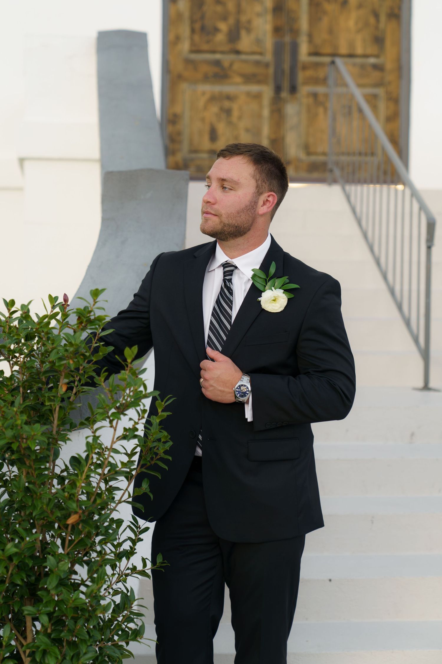 Dapper groom in black suit with white boutonniere stands confidently outside white church entrance.