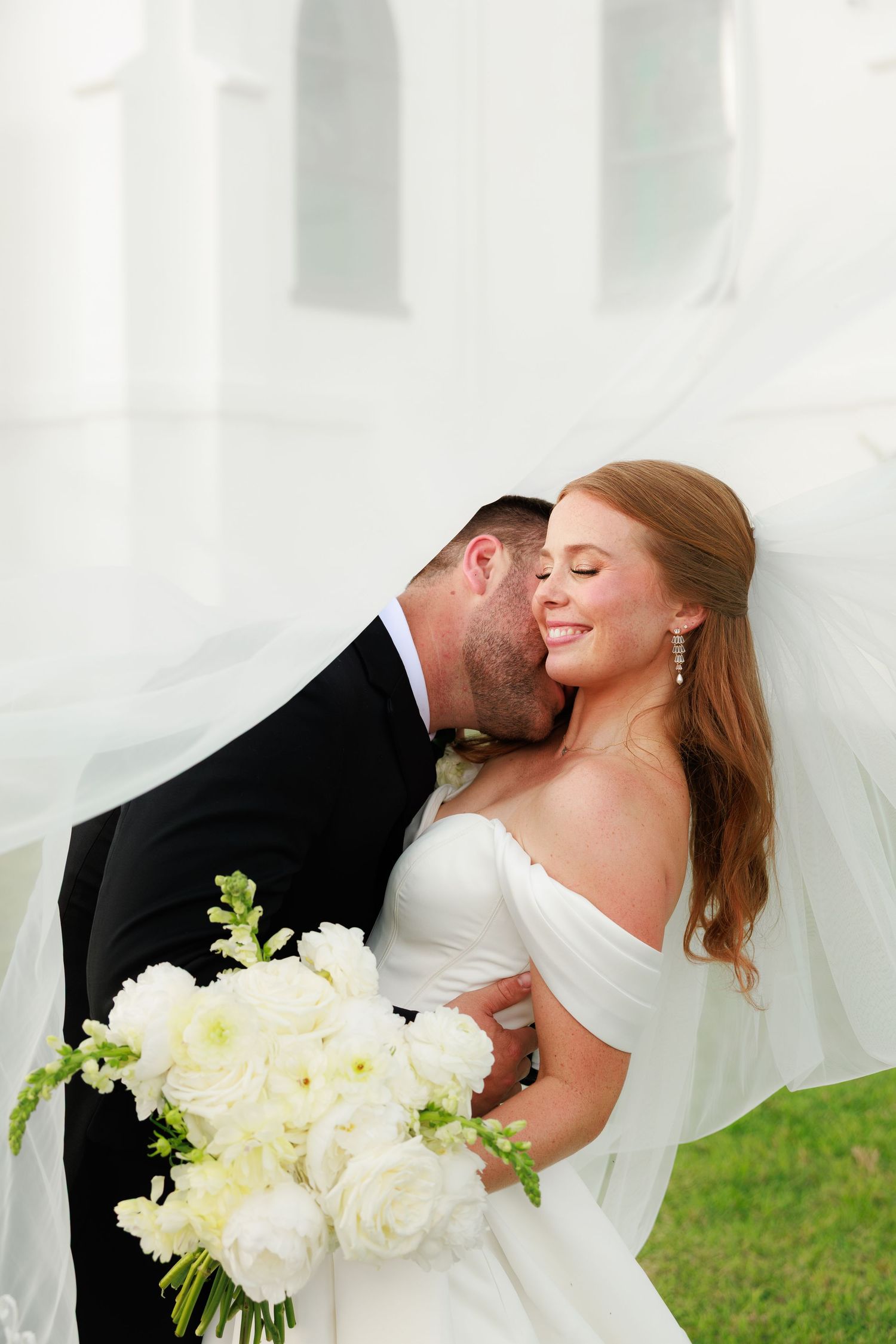 Groom kisses smiling bride on cheek as flowing veil surrounds them, bride holding white rose bouquet.