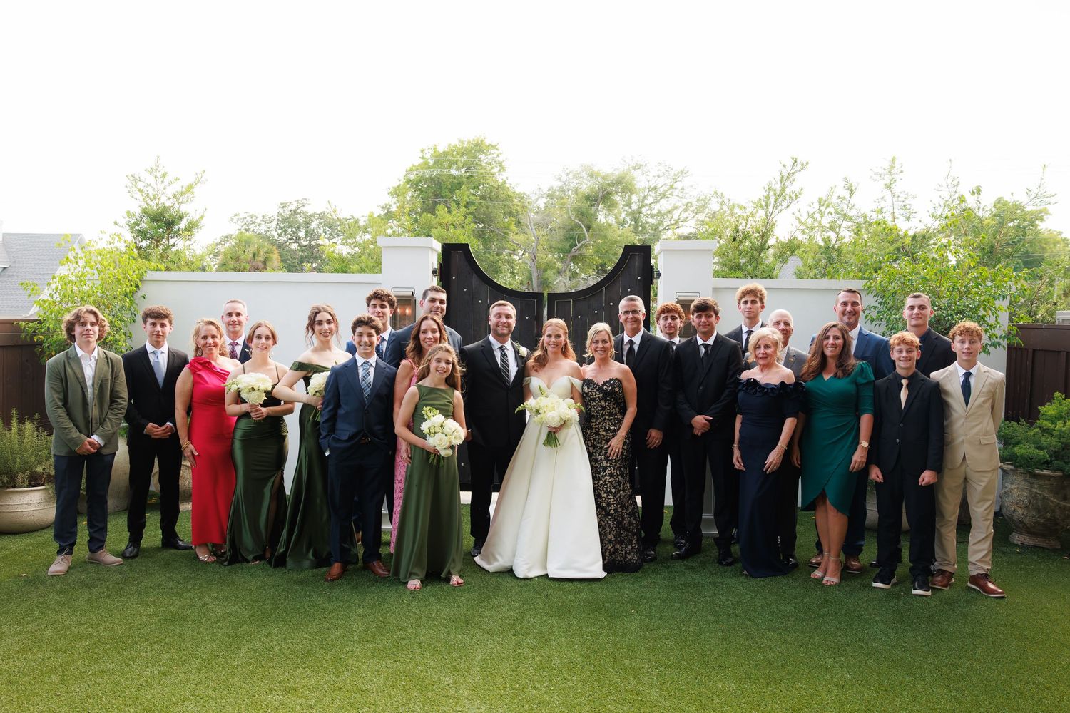 Large wedding party poses outdoors on green lawn with bride and groom at center, guests in formal attire.
