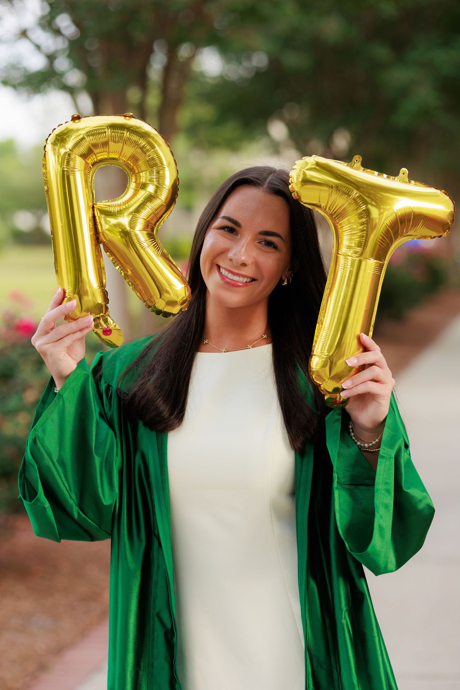 A smiling graduate in a green cap and gown holds gold letter R and T balloons outdoors.