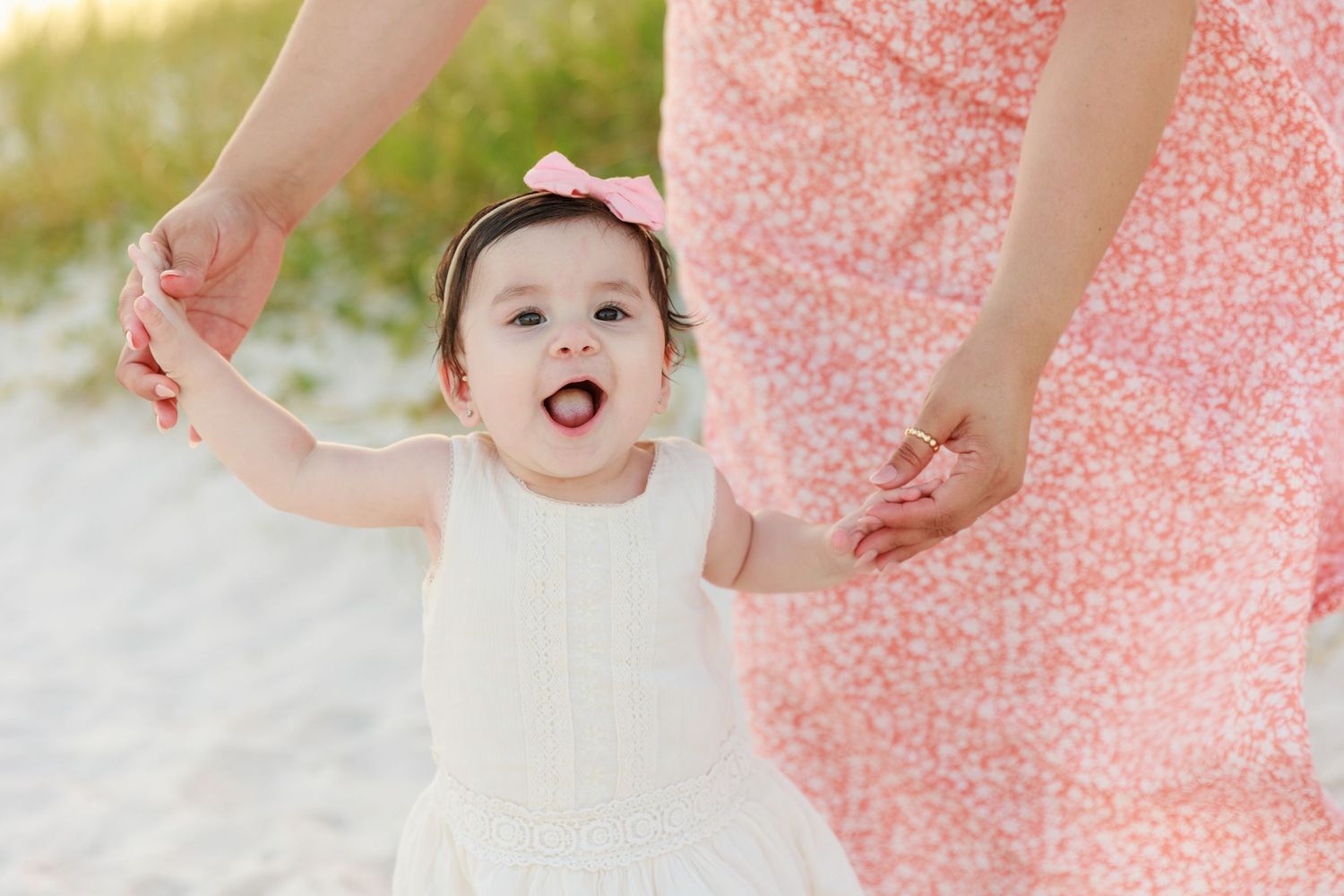 Joyful baby girl in white dress holds hands with mother in floral pink dress on sandy beach.