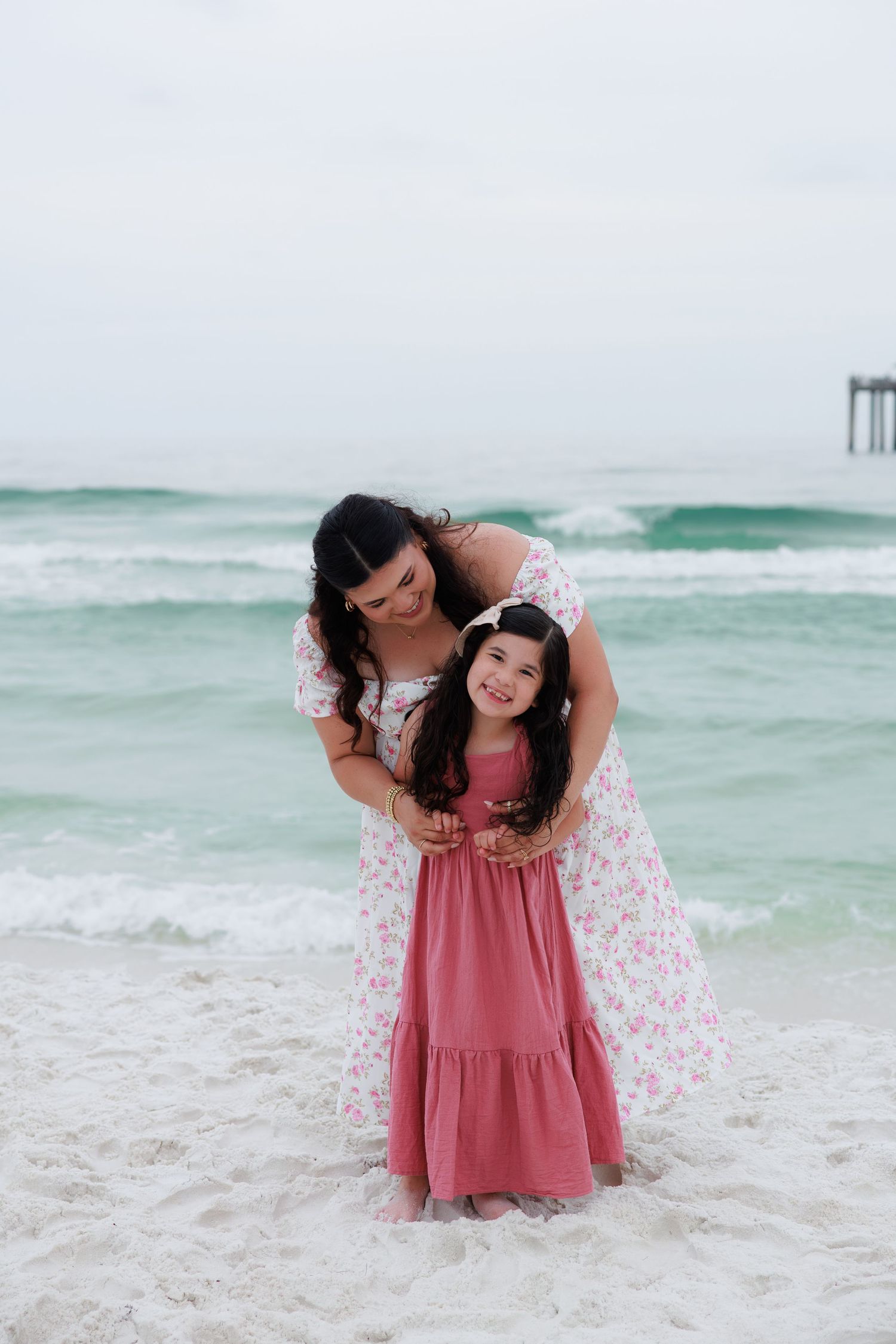 A mother and daughter in matching pink dresses smile and embrace on a white sandy beach with turquoise ocean waves.