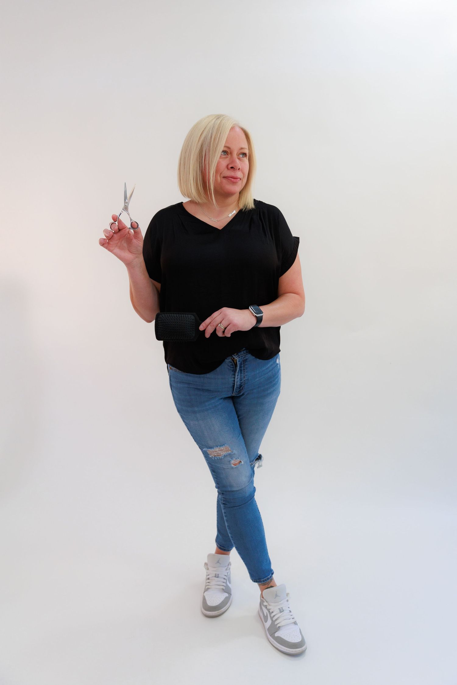 Professional wearing black v-neck top and distressed jeans poses casually against white backdrop with scissors.