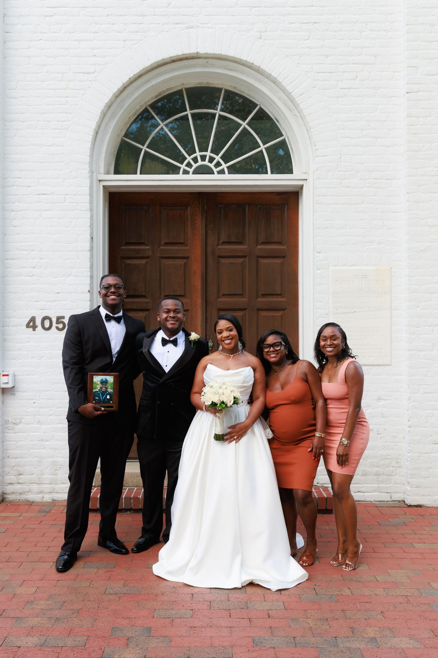Wedding group gathers for formal photos in front of a white church building with arched windows.