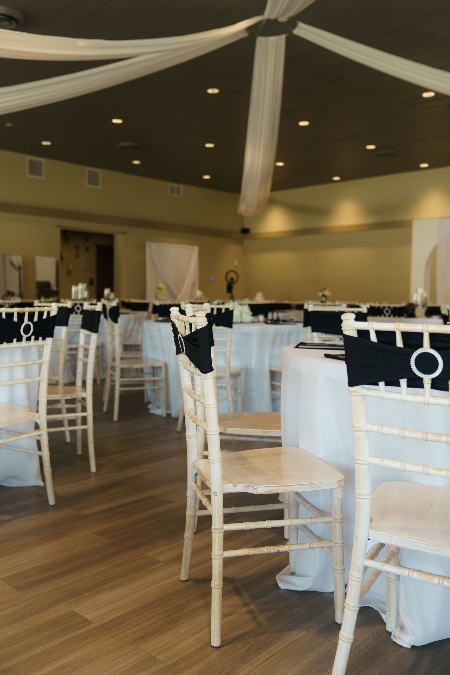 White chiavari chairs arranged in rows on a hardwood floor in an elegant event venue with draped fabric.