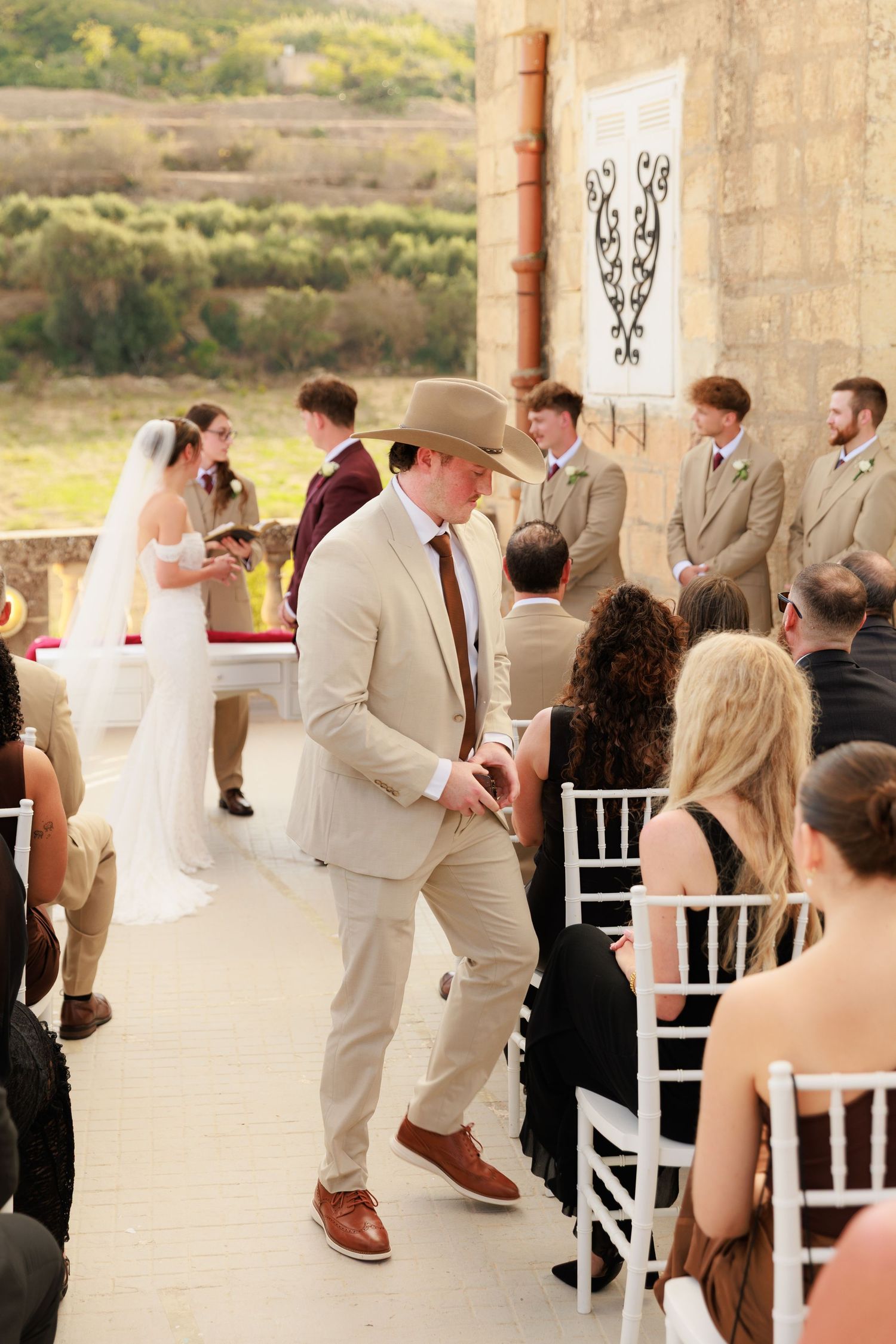 Wedding guests perform a choreographed dance sequence at an outdoor venue with stone columns and scenic views.