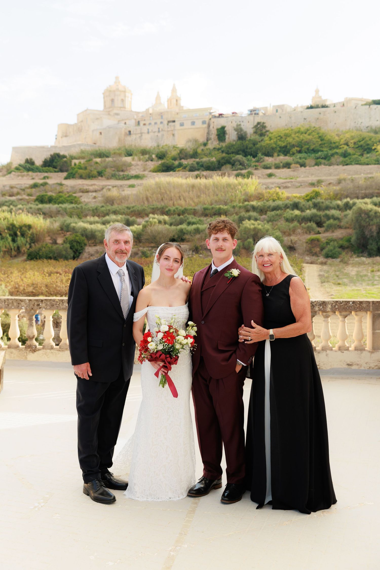 A wedding party poses outdoors on a stone terrace with ancient walls and Mediterranean scenery in the background.