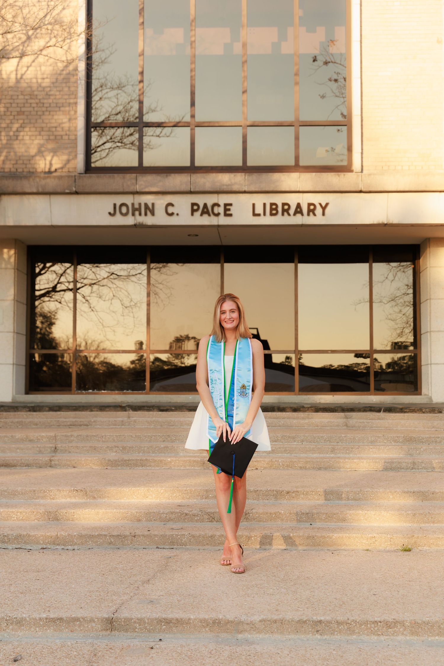 Graduate in cap and gown poses on steps outside John C. Pace Library wearing light blue graduation stole.