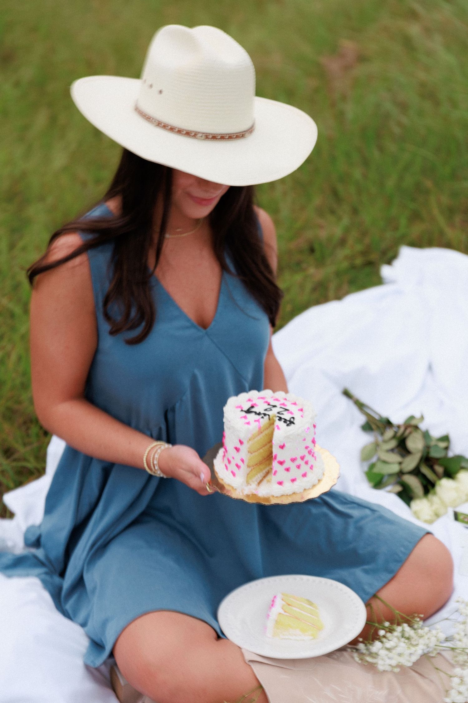 Person in blue dress and white hat holding small decorated cake during outdoor picnic.
