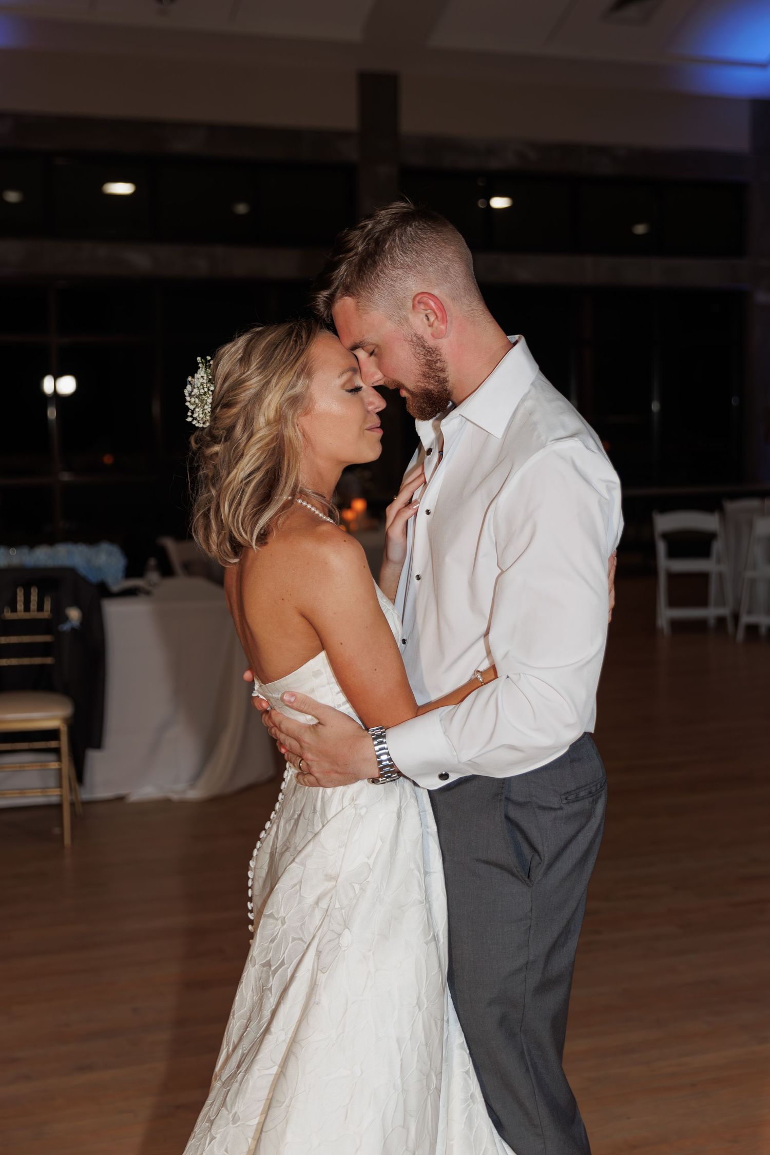 Newlyweds share intimate first dance moment in reception hall with white chairs visible.