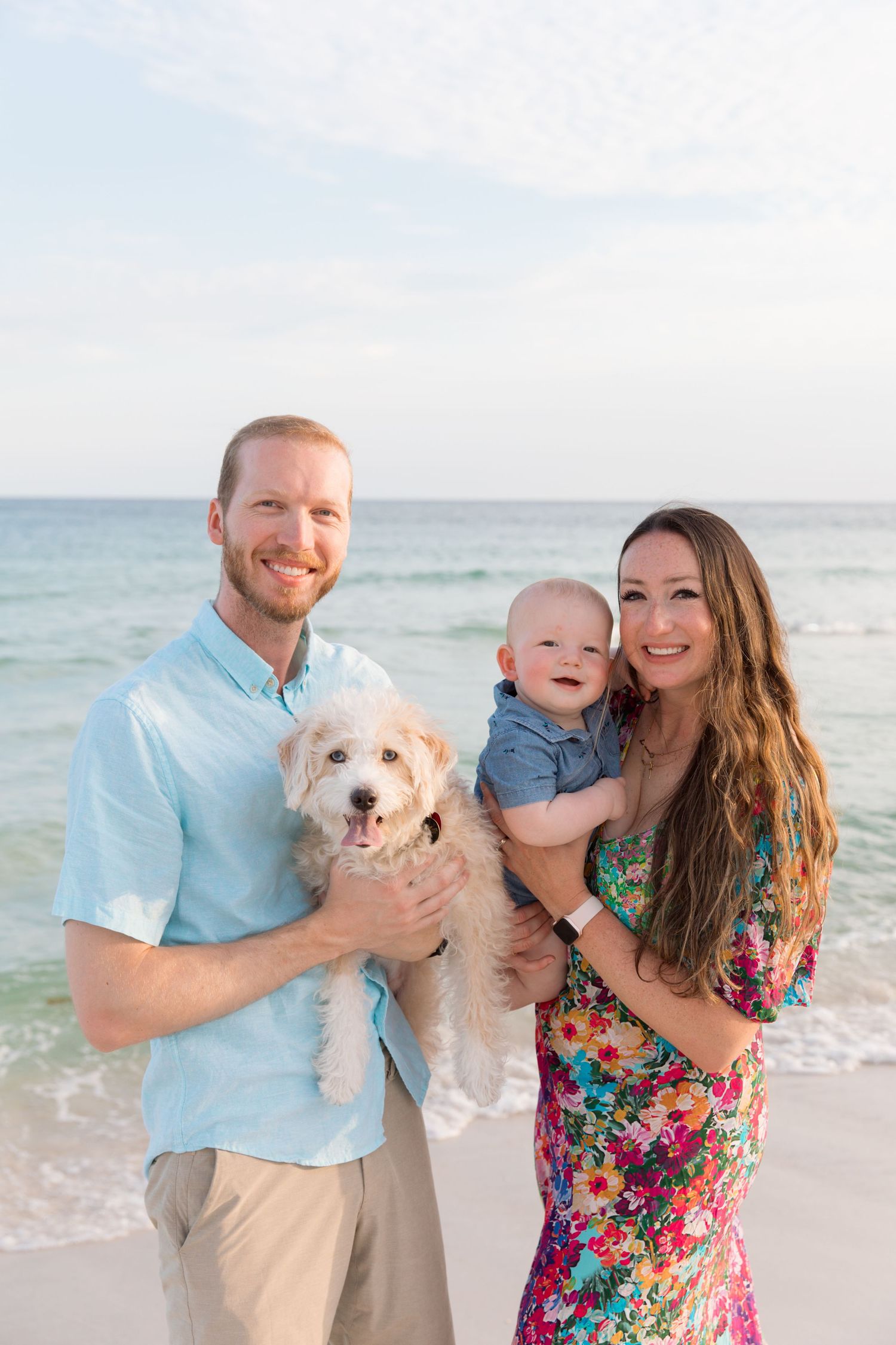 A happy family poses for beach portraits with their white fluffy dog by the ocean.