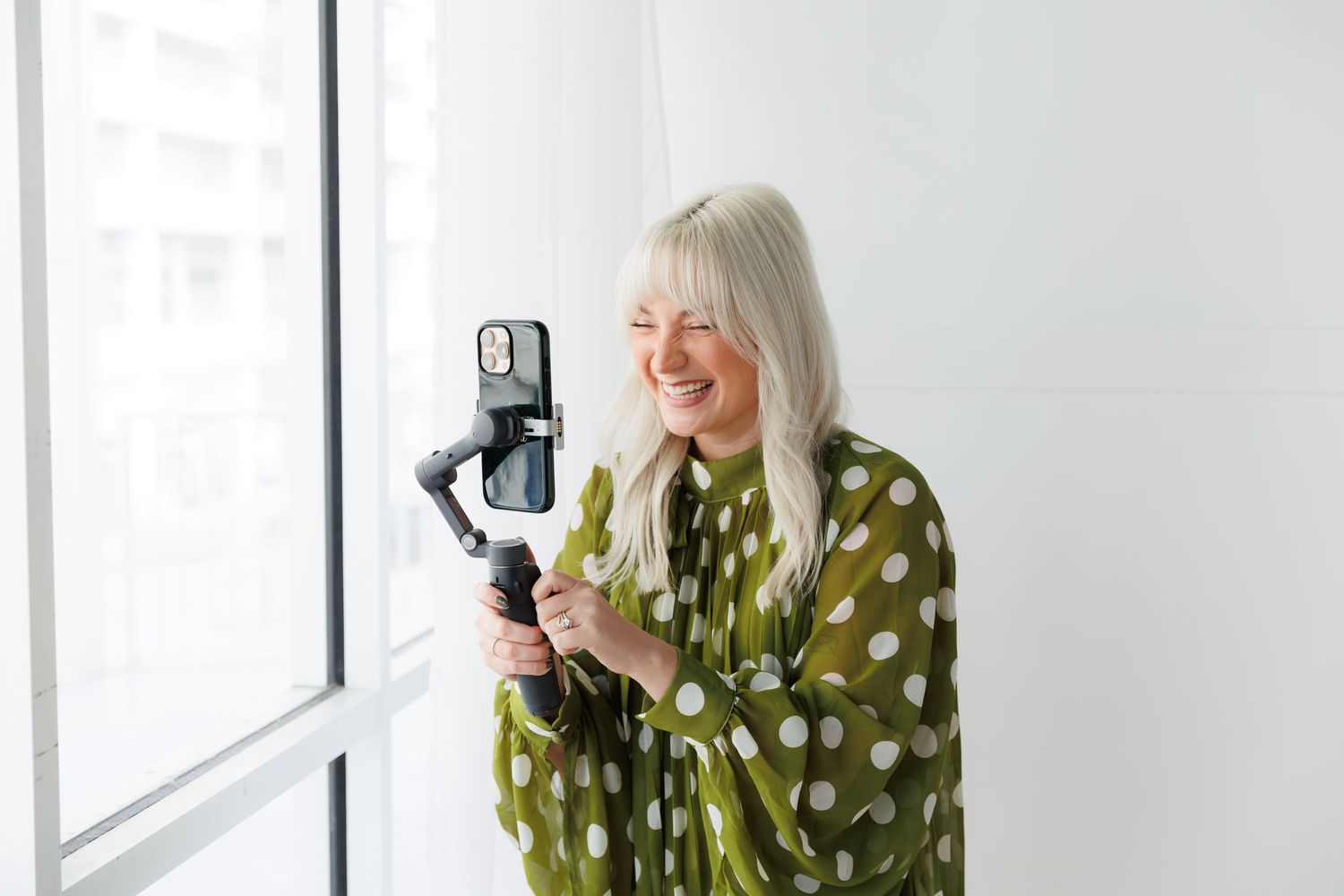 Woman in green polka dot dress poses near window with phone and ring light for content creation.