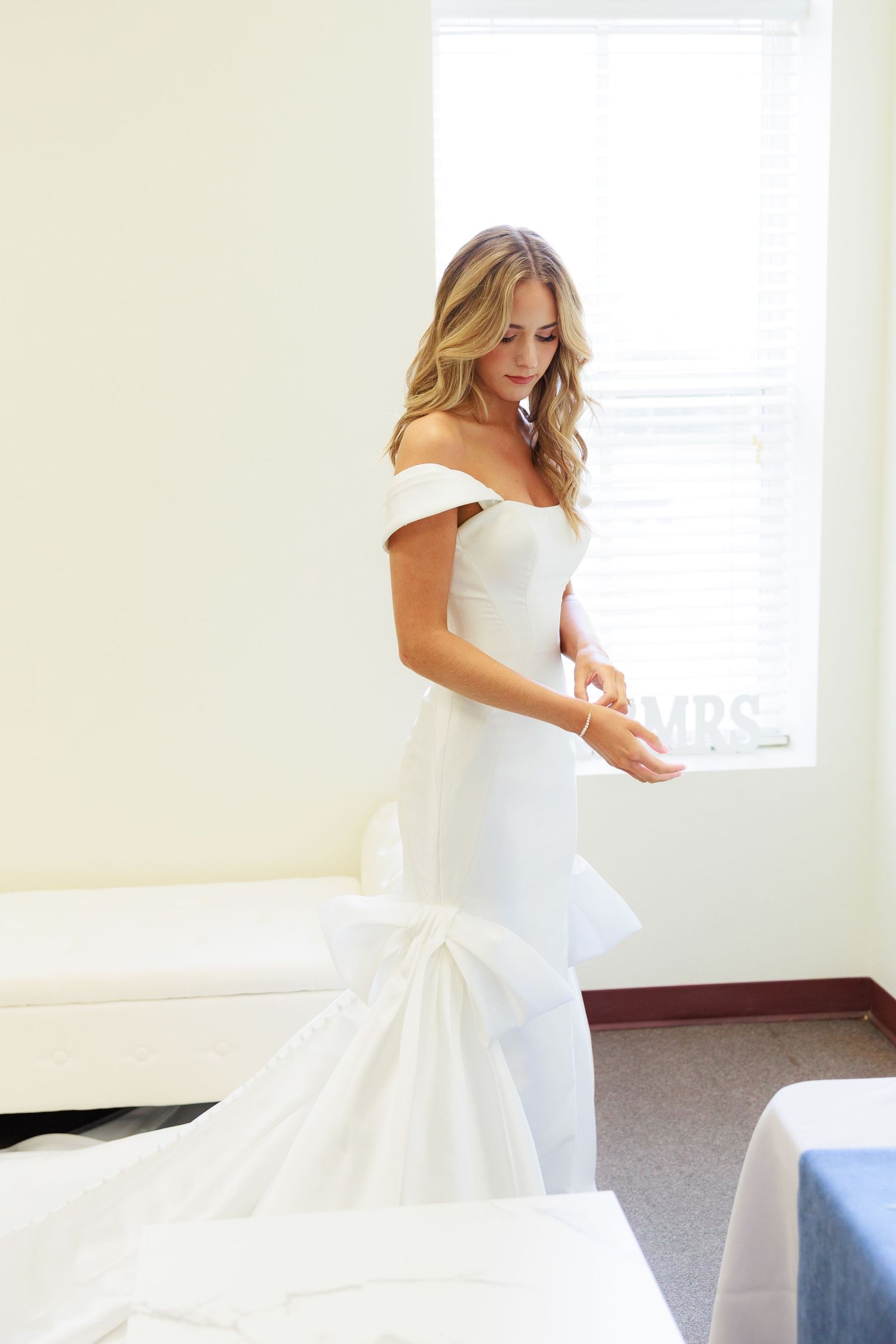 A wedding dress with off-shoulder design and mermaid silhouette is displayed in a bright room.