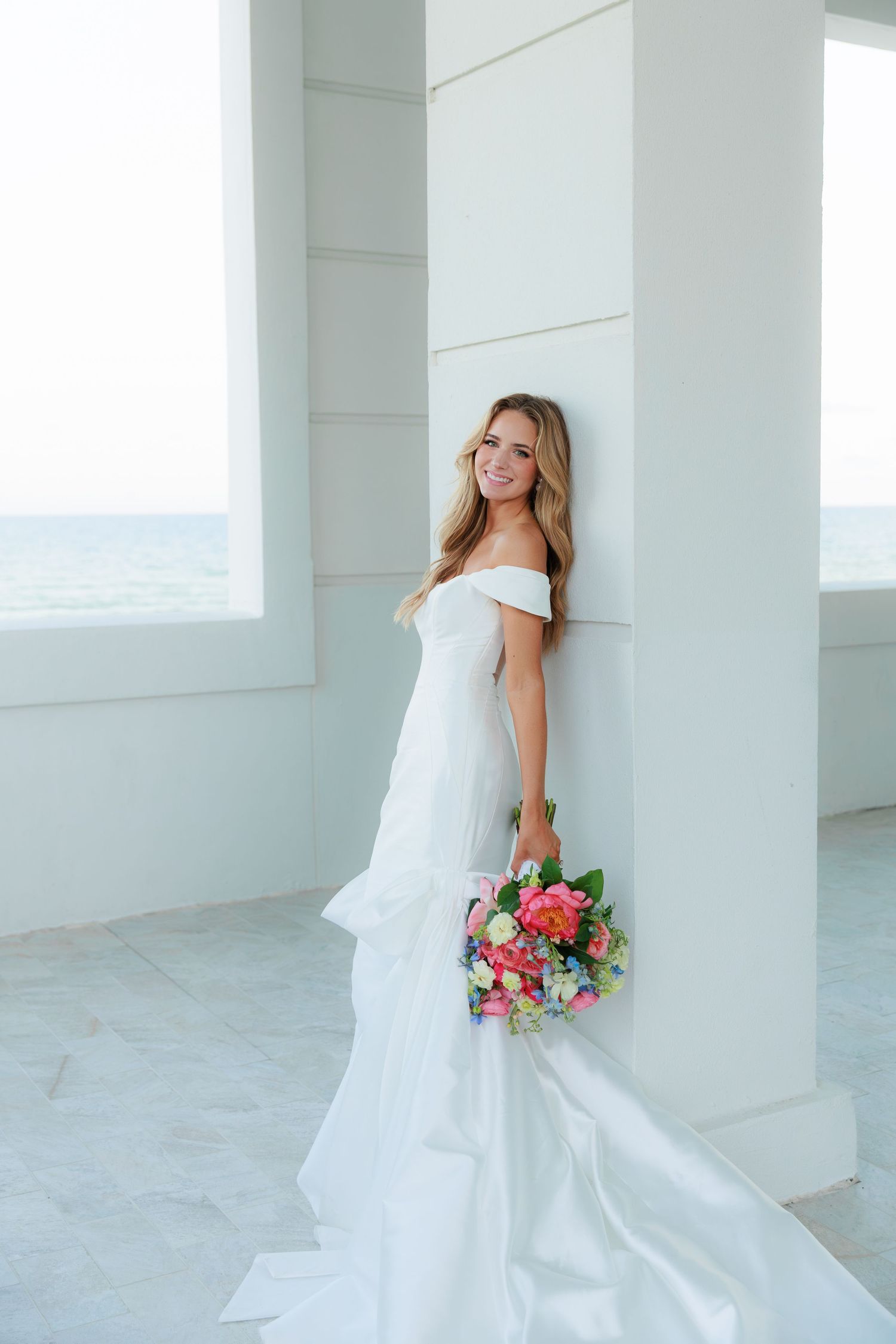 Elegant bride in white off-shoulder gown poses with vibrant bouquet against modern white architecture.