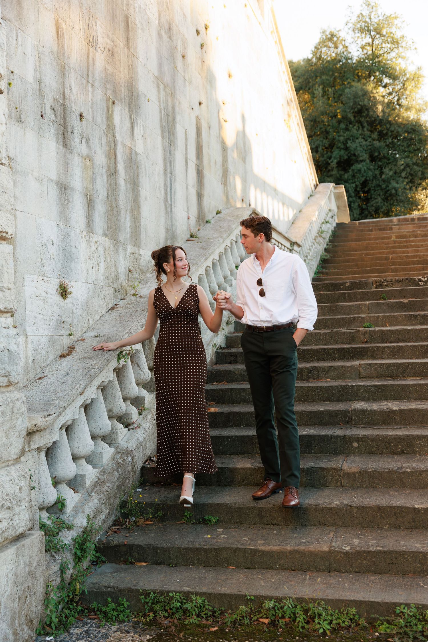 Elegant couple holding hands on stone steps, woman in polka dot dress, man in white shirt and dark trousers.
